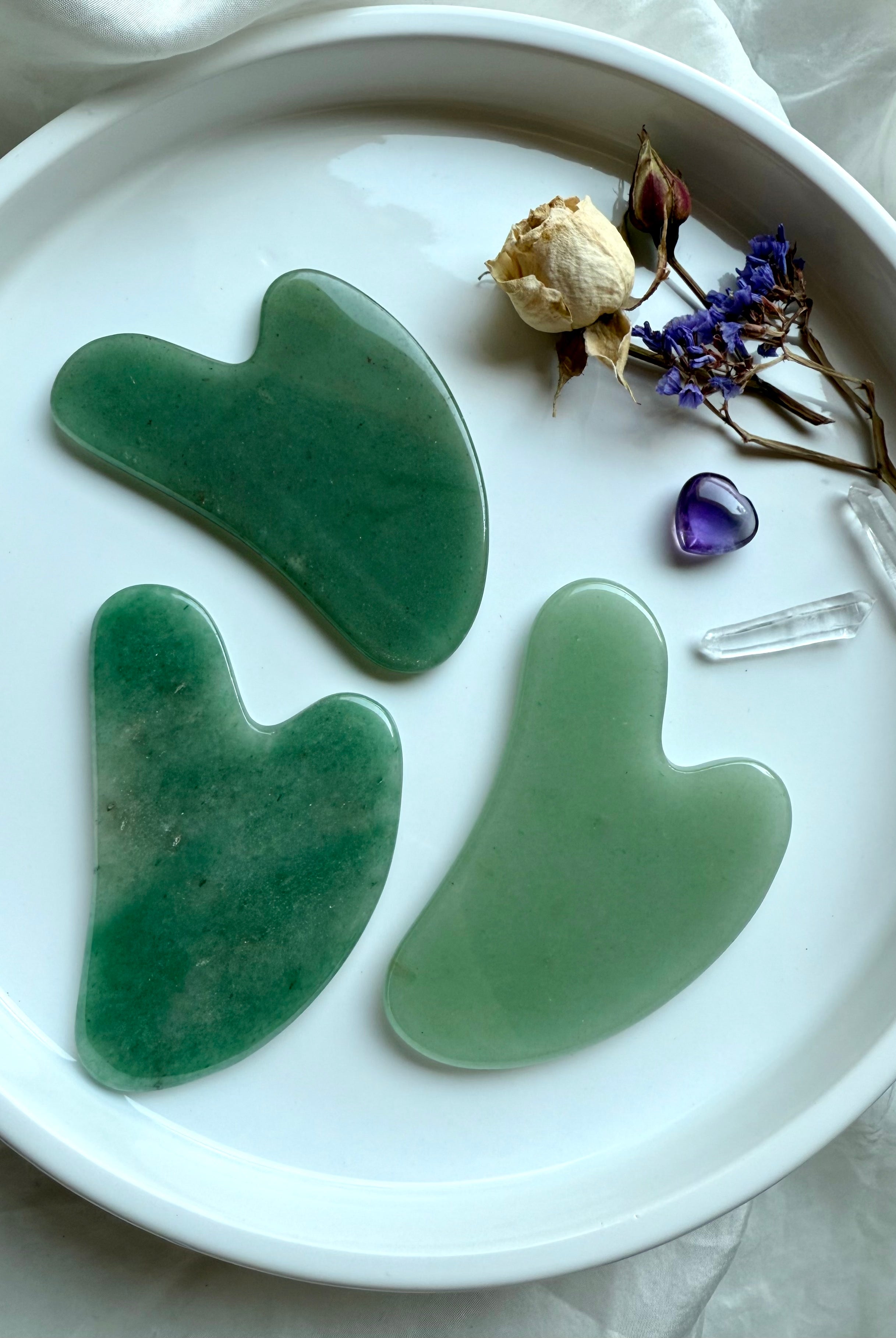 Set of three Green Aventurine crystal gua sha facial massage tools on white plate with floral accents and quartz points