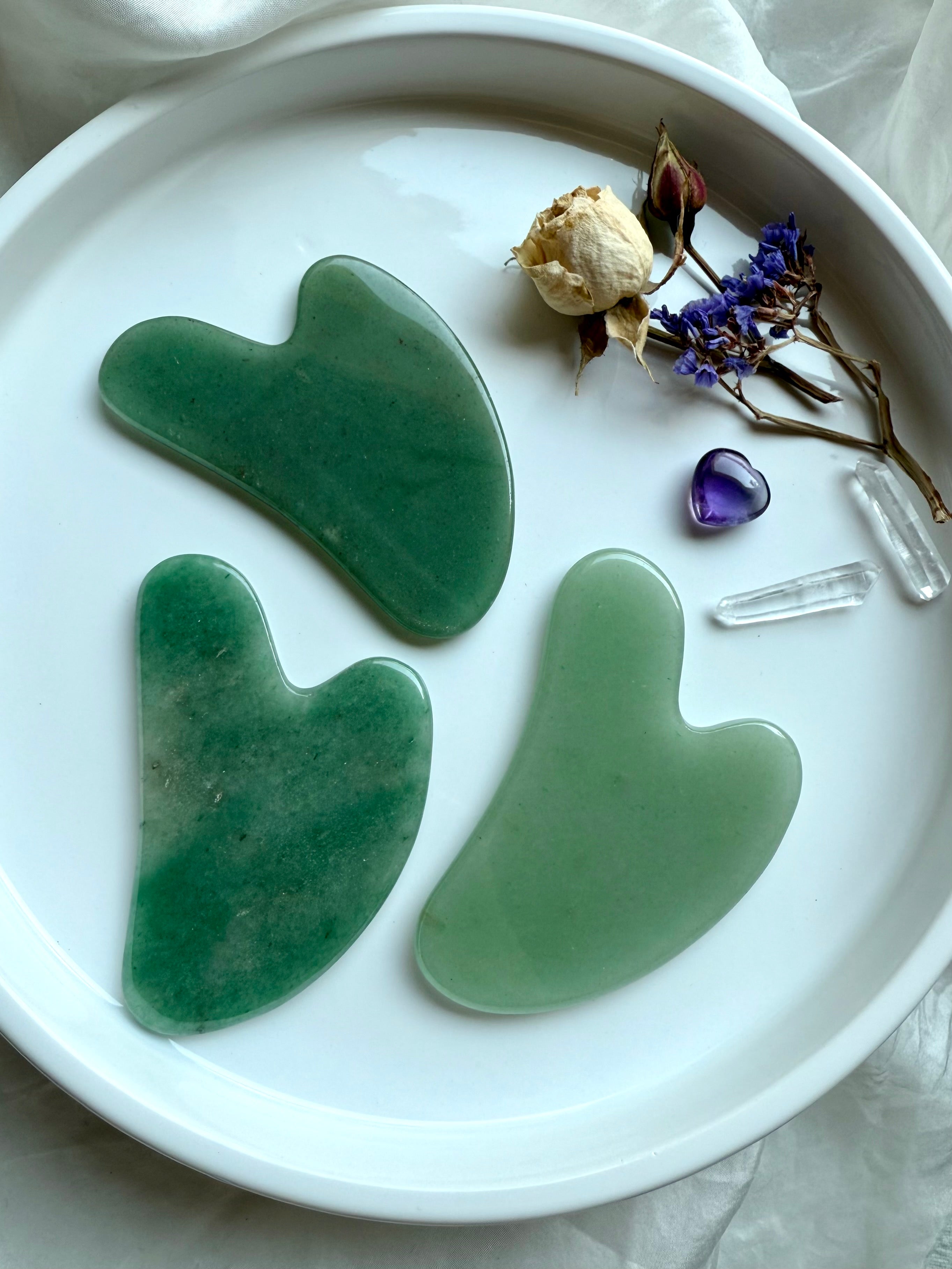 Set of three Green Aventurine crystal gua sha facial massage tools on white plate with floral accents and quartz points