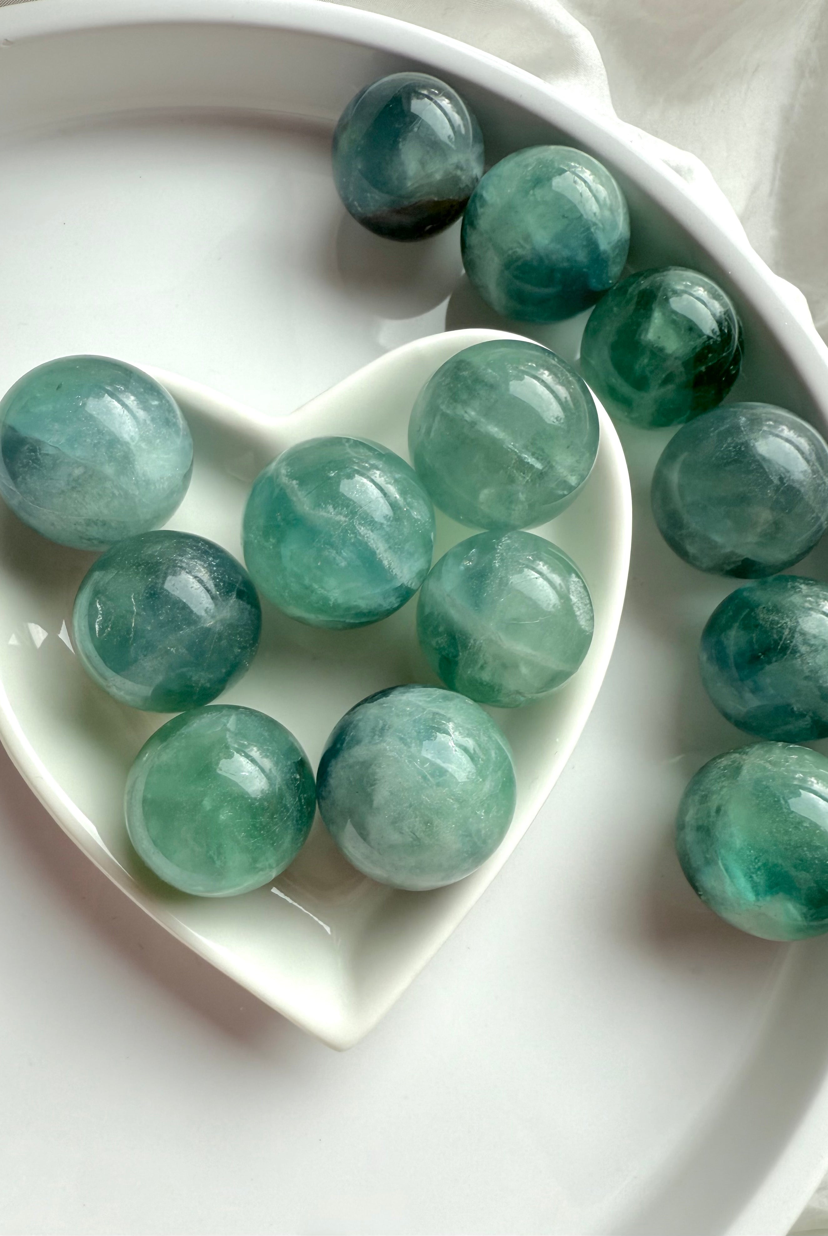 Mini teal Fluorite spheres in a white heart dish, radiant aqua blue crystal balls for meditation and chakra energy work.
