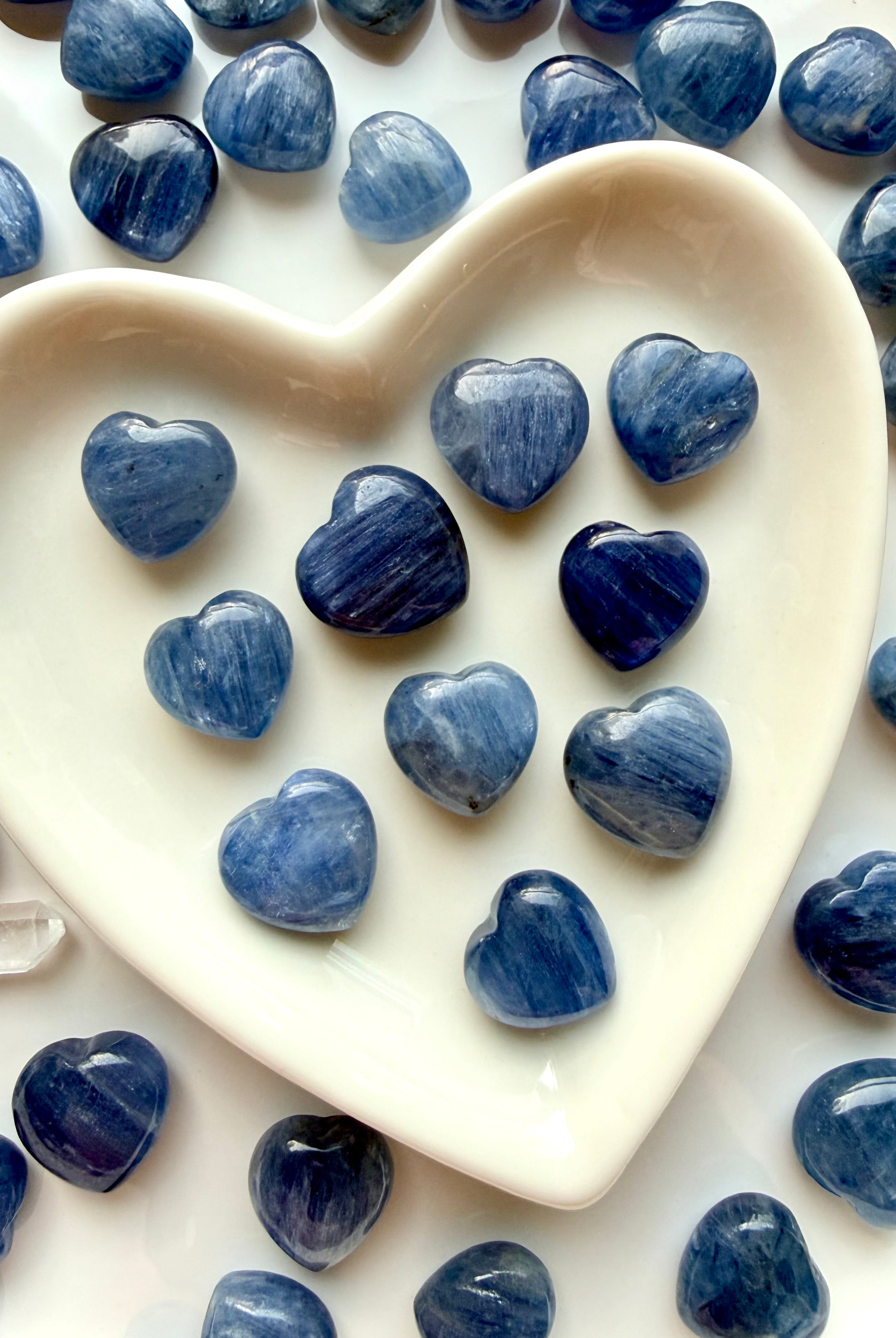 Mini Blue Kyanite heart crystals displayed in a white heart bowl with additional hearts in the background, throat chakra stones for communication and calm