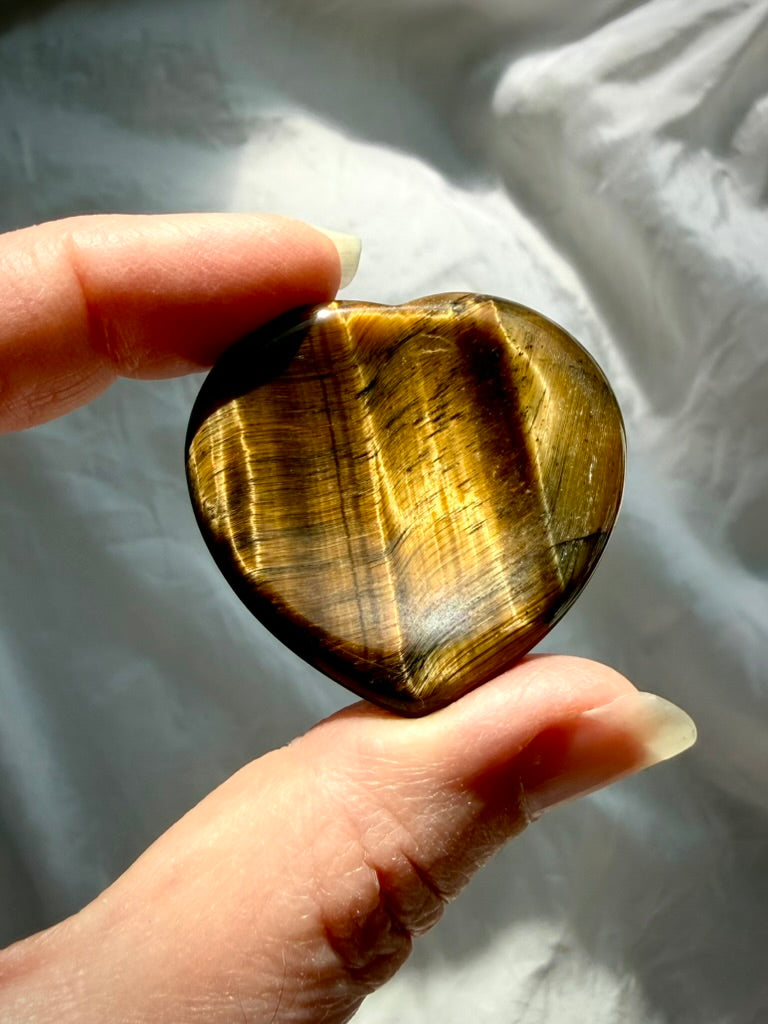 Polished Tiger’s Eye heart worry stone held in sunlight, displaying luminous golden flash and natural mineral banding