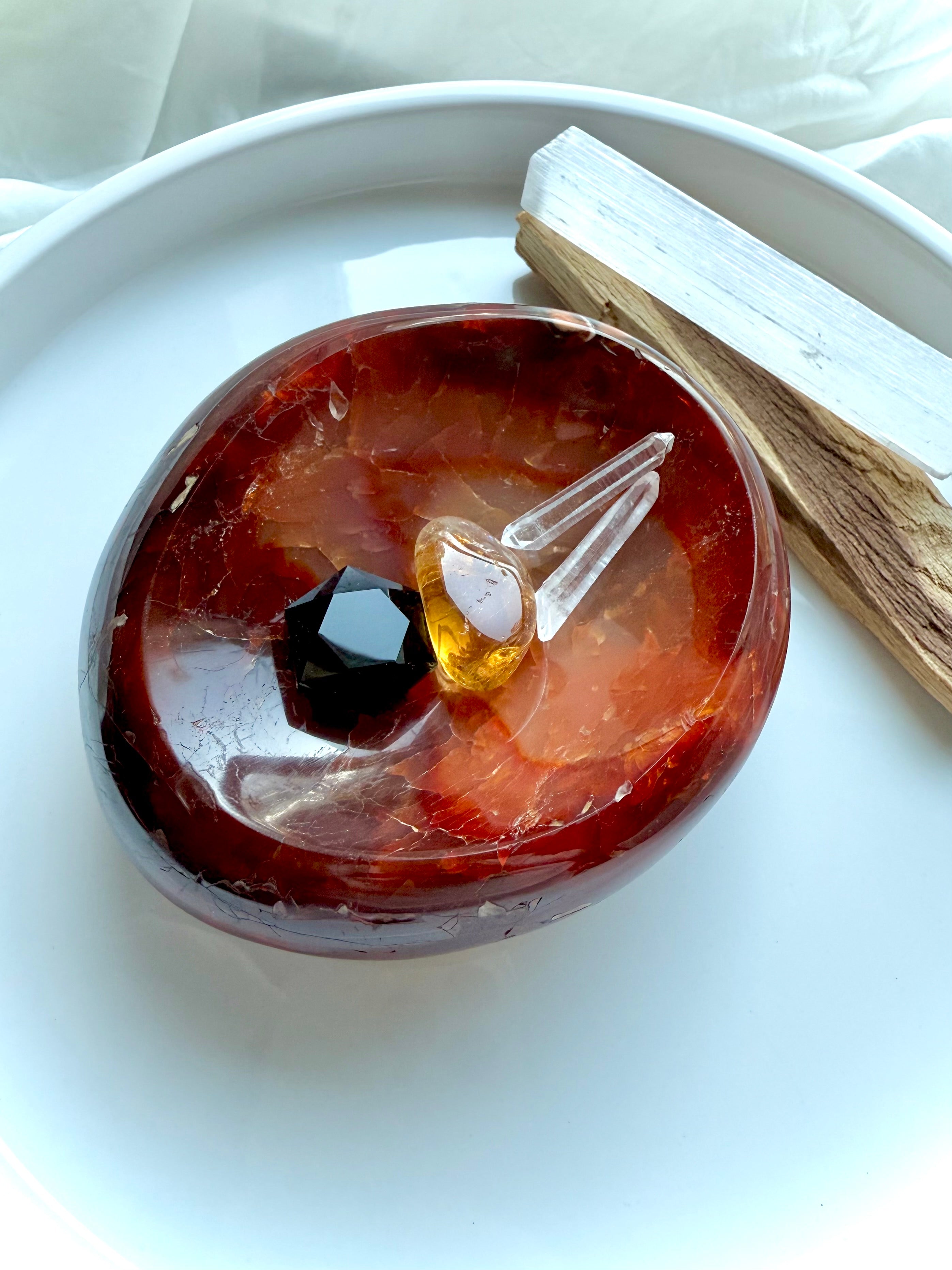 Carnelian crystal bowl displayed with a Clear Quartz point, citrine tumble, Black Obsidian gem, Selenite stick, and Palo Santo on a white ceramic plate – ideal for altar or energy cleansing.