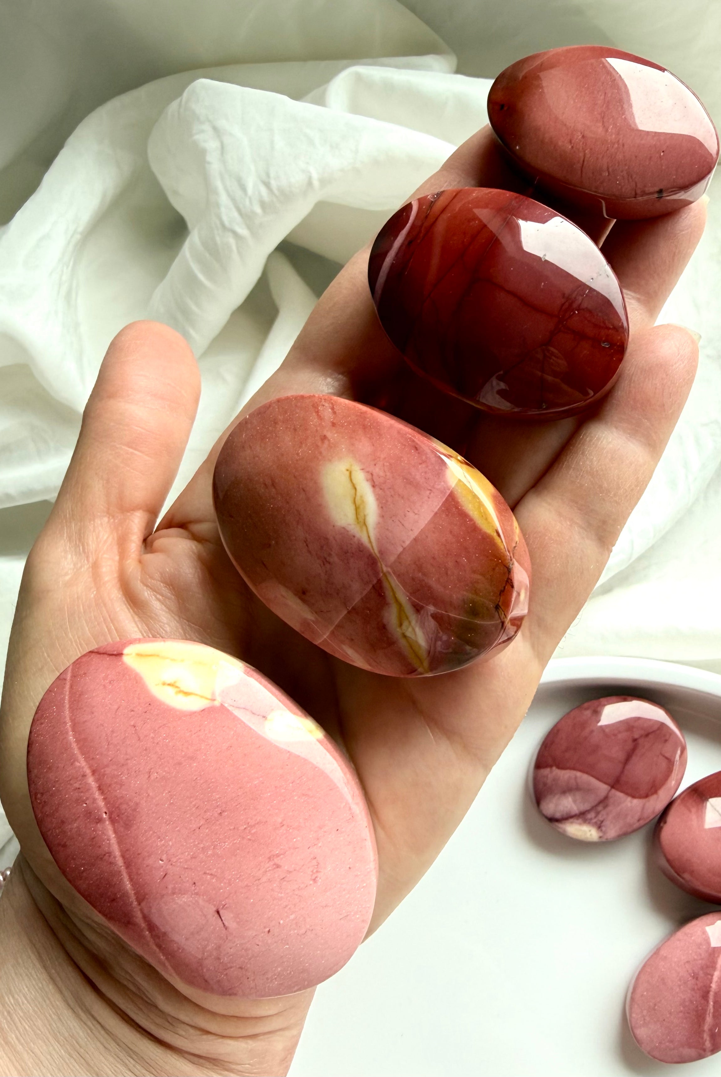 hand holding smooth Mookaite Jasper palm stones with natural pink, cream, and deep red banding for grounding energy