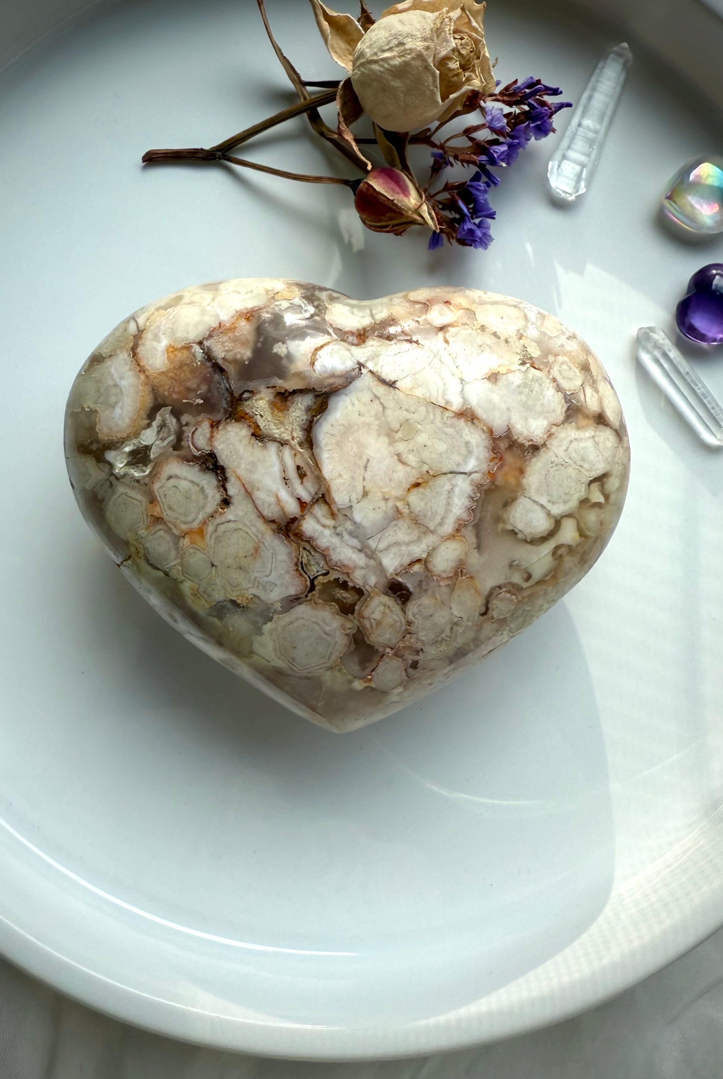 Polished Flower Agate heart showing natural flower-like formations in beige and soft blush tones, styled with dried roses and aura accents — nurturing crystal for emotional healing and gentle transformation.