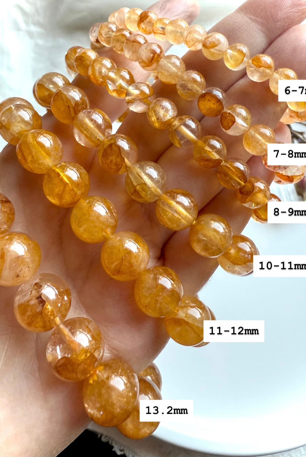 Golden Healer Quartz bracelet stack on hand with bead sizes labeled 6–7mm through 13.2mm, showing variation in size and glowing golden hues.