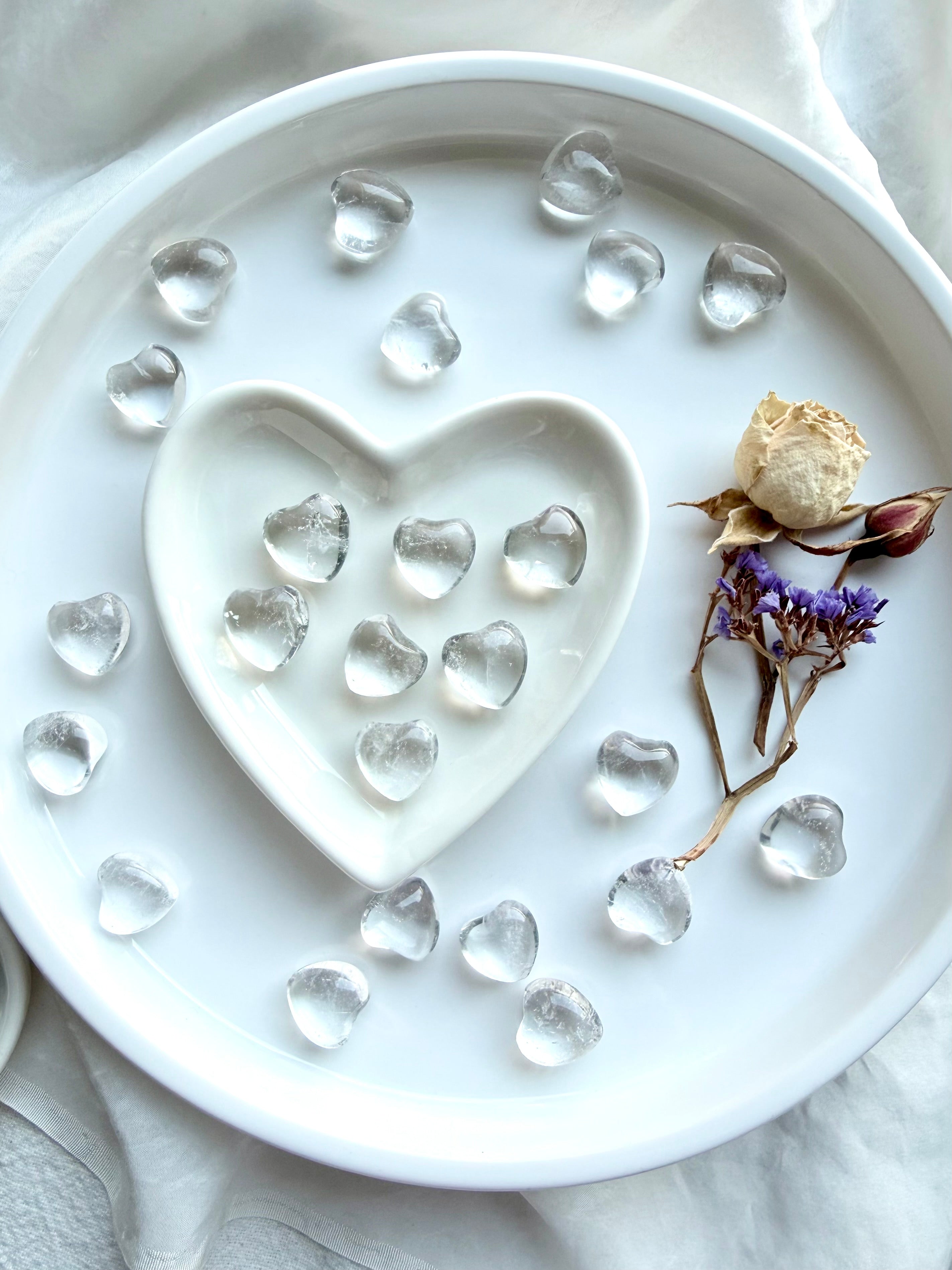 Mini Clear Quartz crystal hearts arranged in white heart bowl with additional hearts on tray with dried flowers
