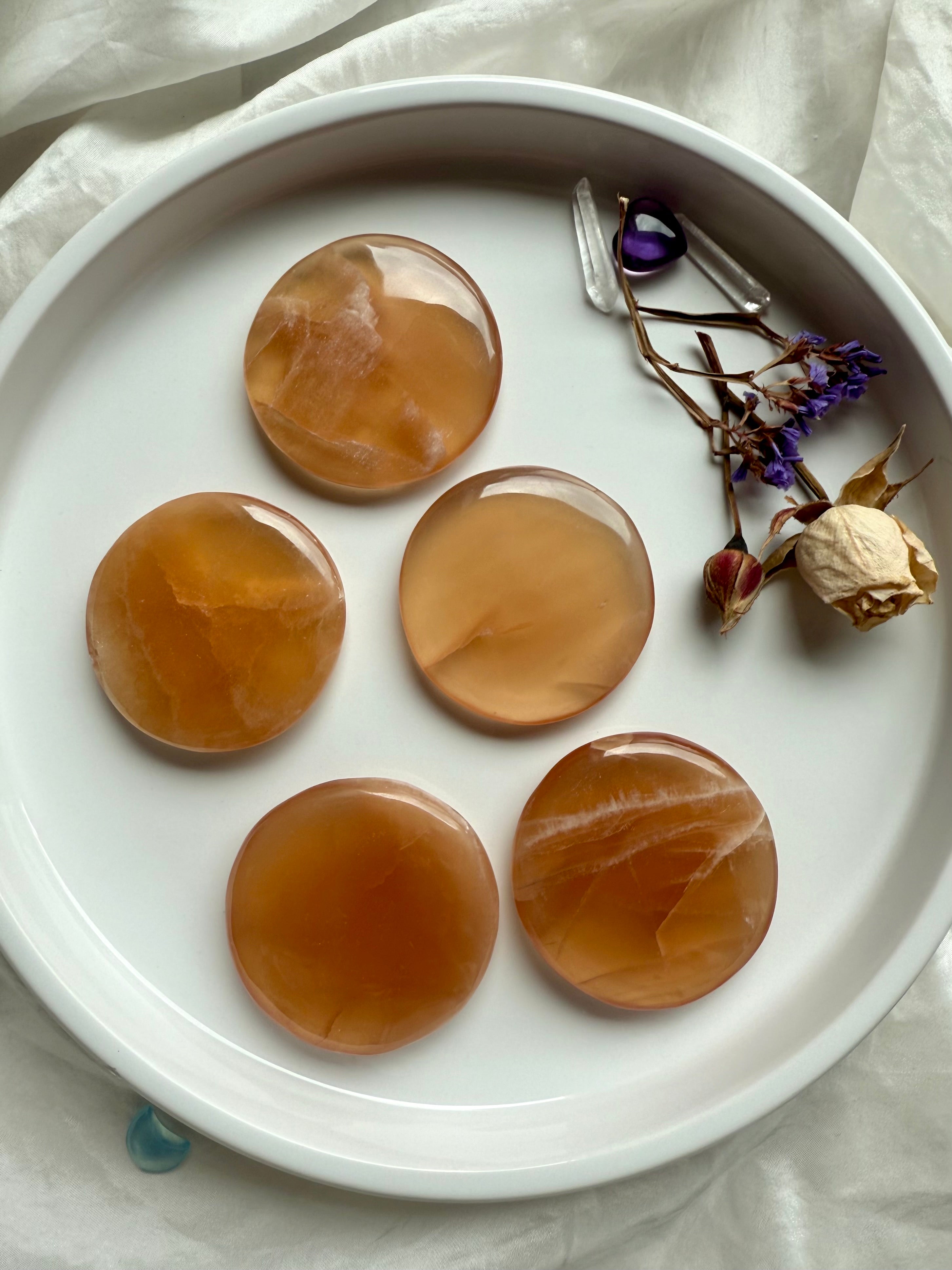 Group of polished honey calcite meditation stones in a white dish with dried flowers, showing soft golden tones and natural banding.