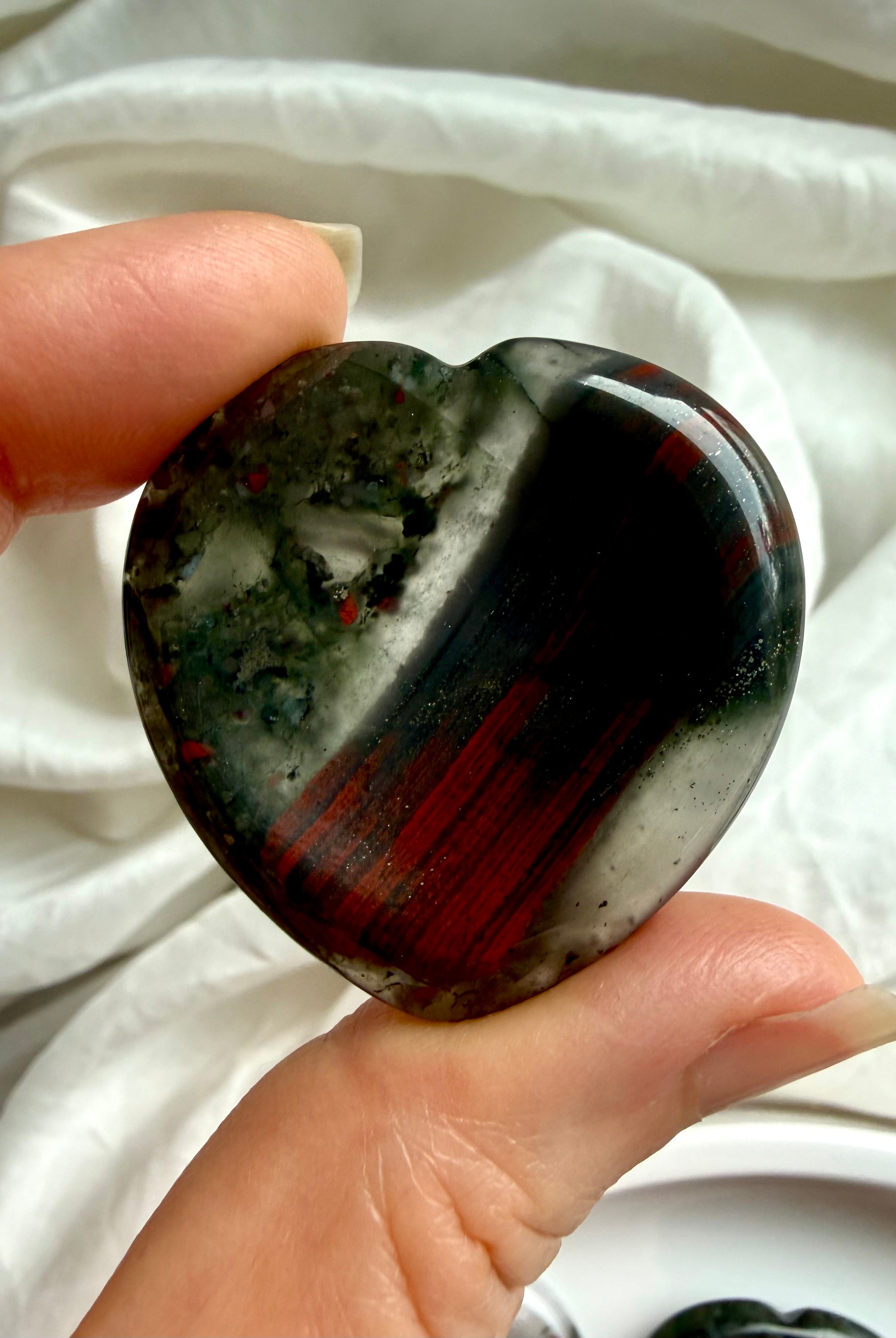 close-up bloodstone heart worry stone held between fingers showing natural pattern variation, crystal for courage stability and stress relief