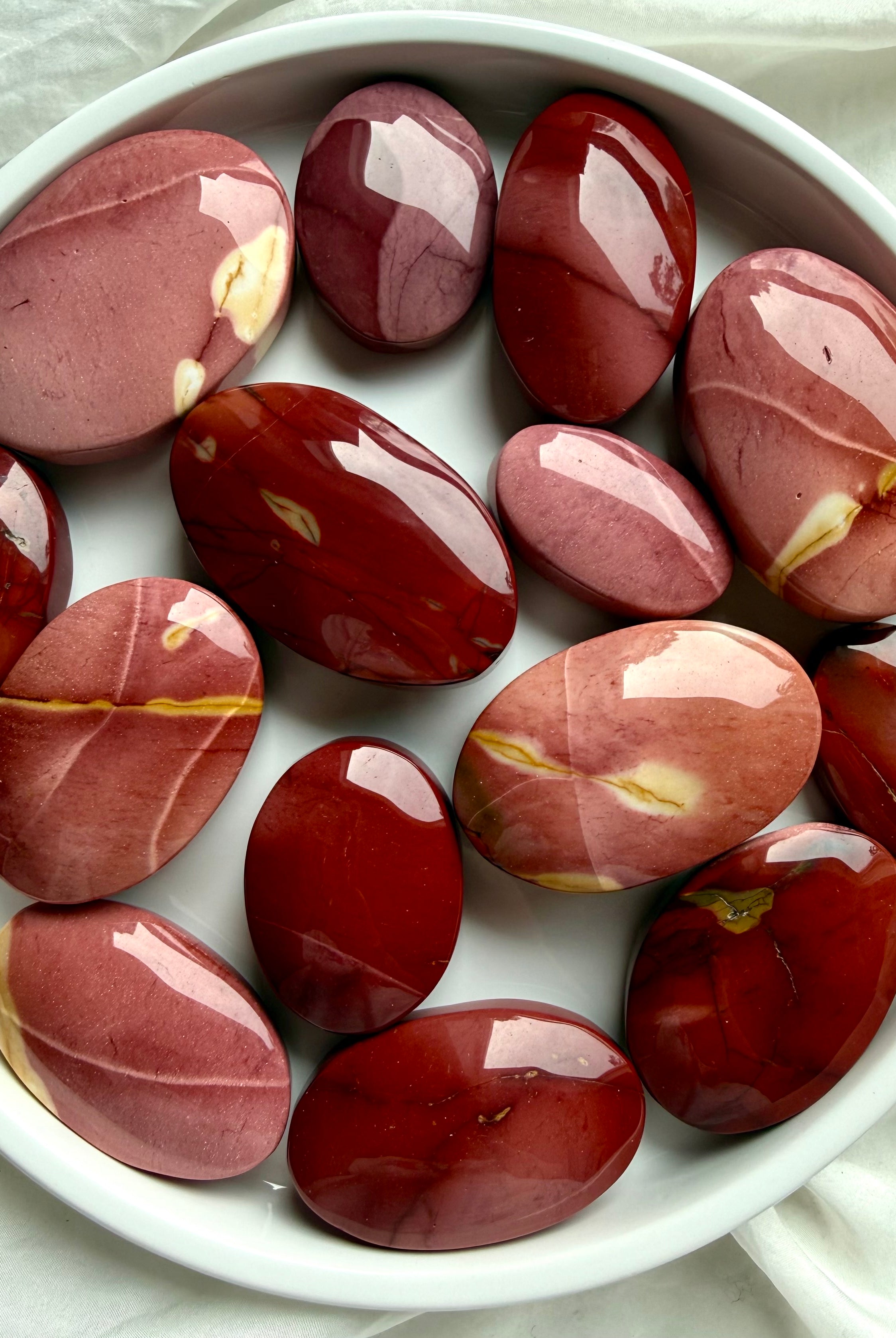 Mookaite Jasper palm stones arranged in white bowl showing natural red, burgundy, and blush color variations