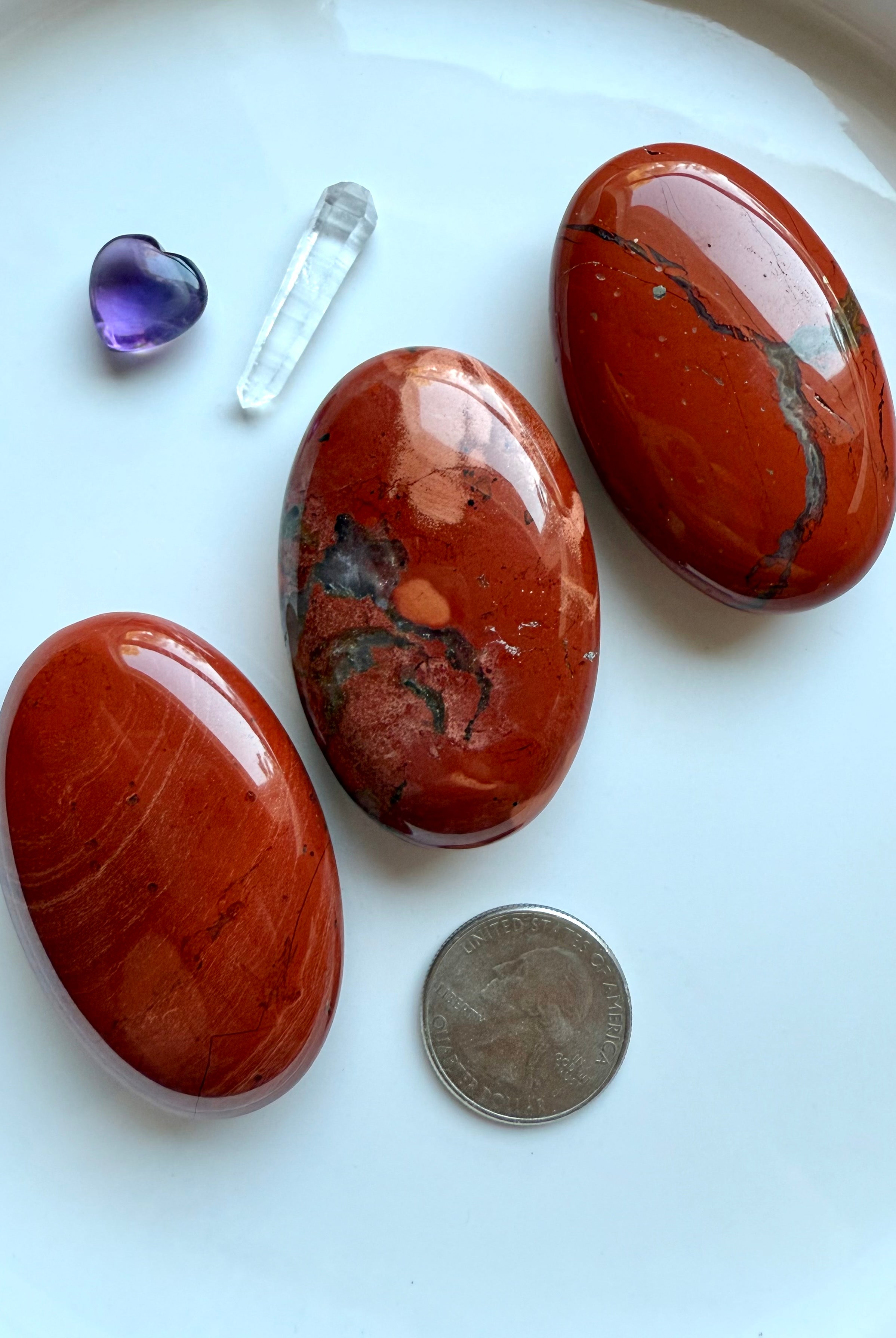 Three Red Jasper palm stones on a white plate with quarter for scale and amethyst heart carving.