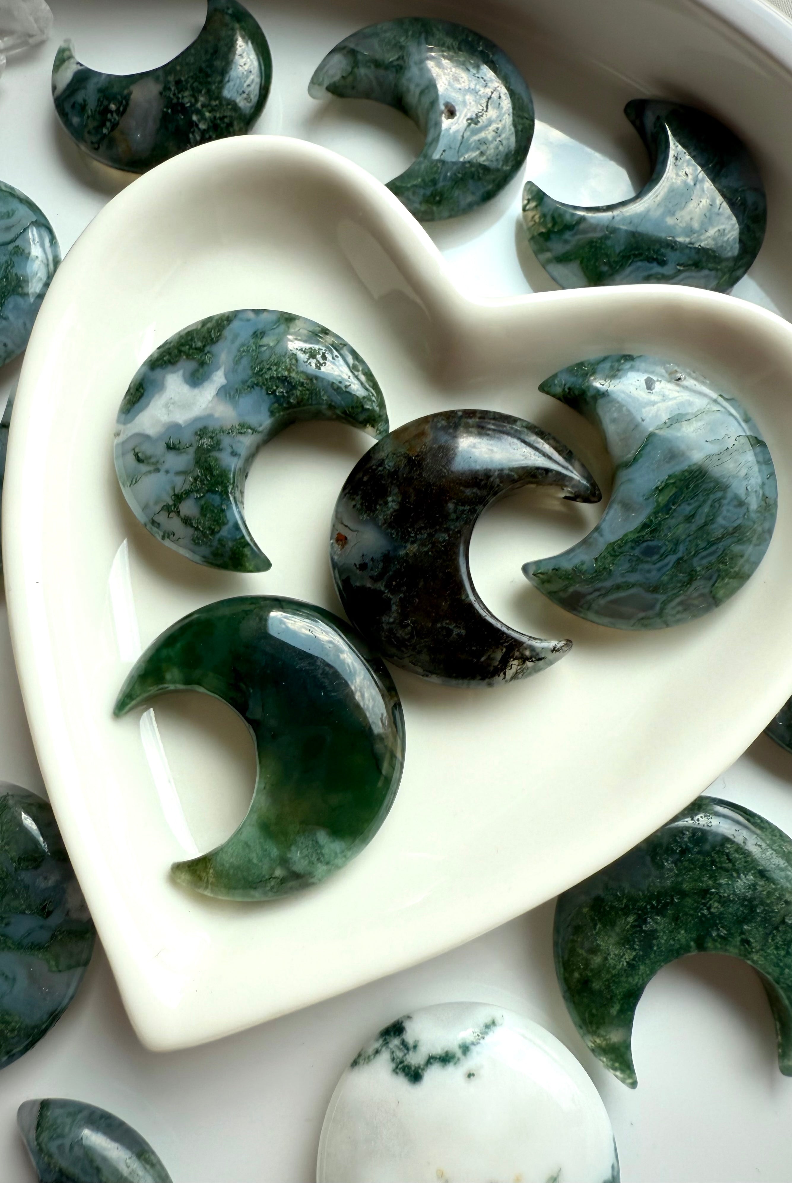 Detailed view of Moss Agate moon carvings in a white heart dish, highlighting natural mossy inclusions and earthy green tones.
