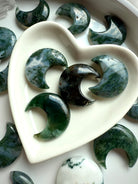 Detailed view of Moss Agate moon carvings in a white heart dish, highlighting natural mossy inclusions and earthy green tones.