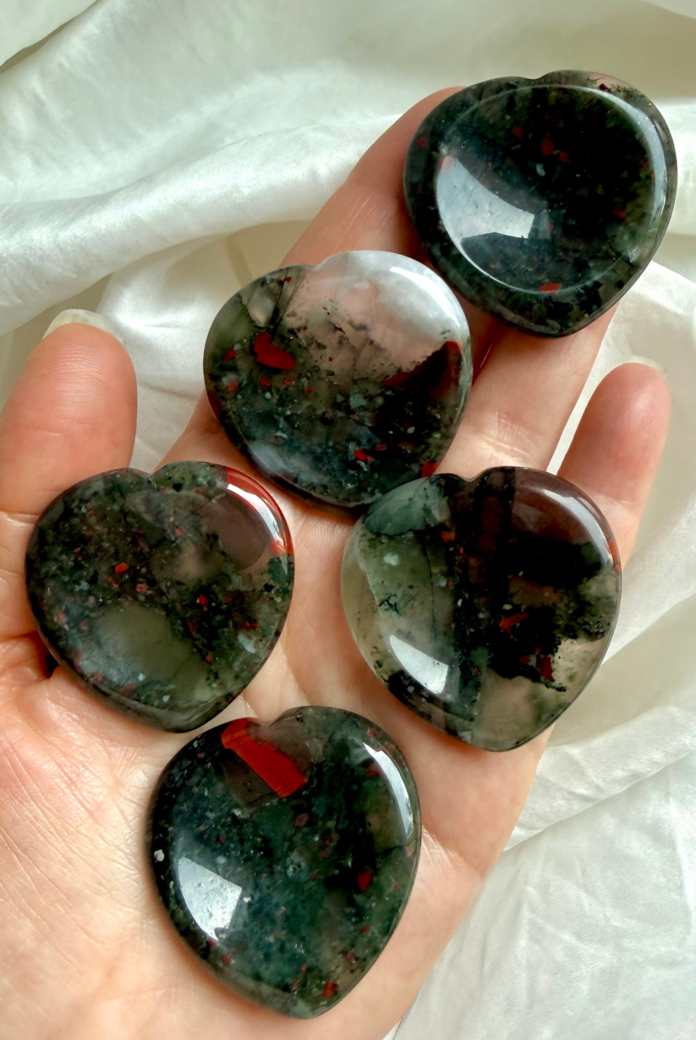 five bloodstone hearts shown on hand with rich green tones and vivid red inclusions, grounding crystal for strength focus and vitality