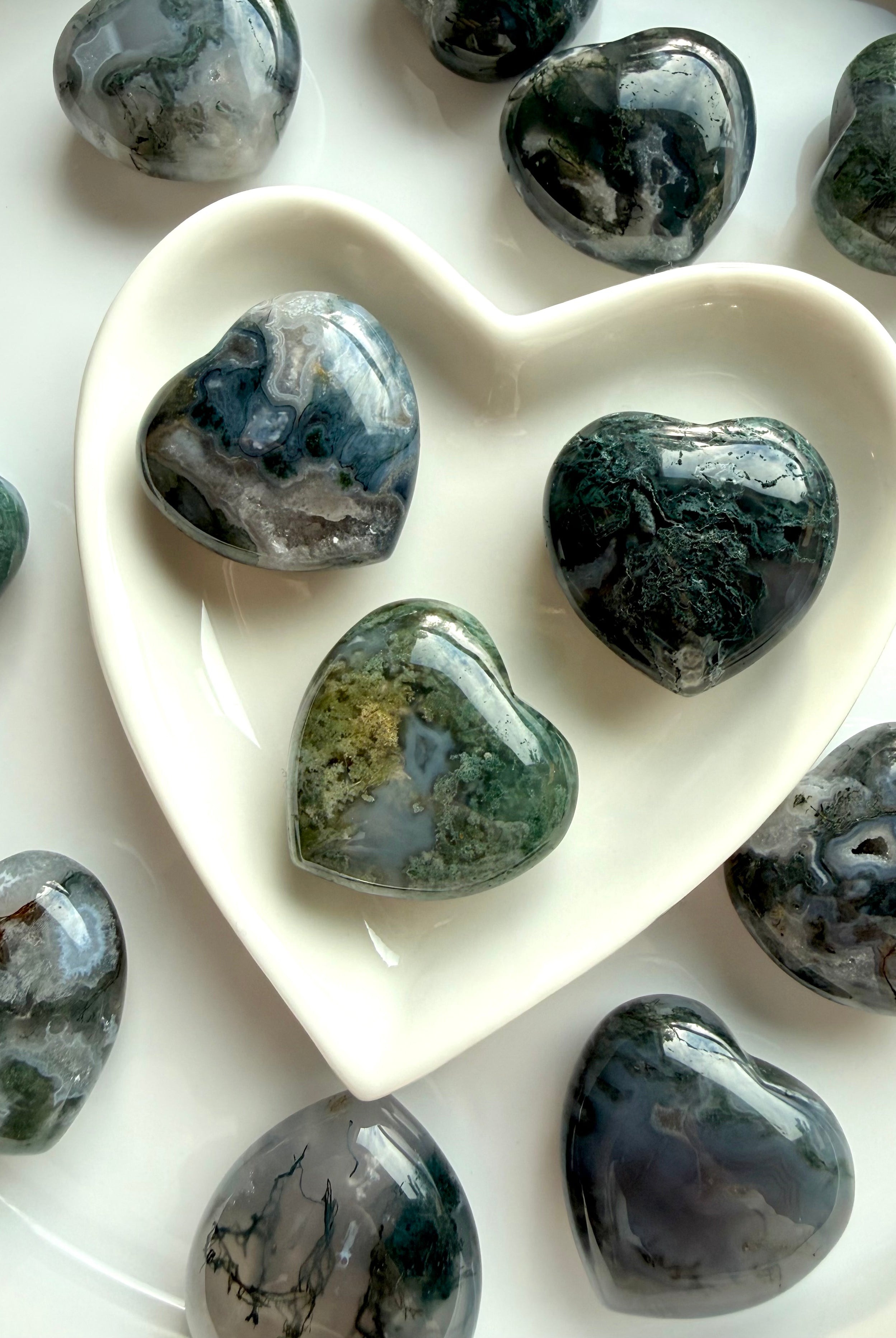 Moss Agate crystal hearts in a white heart-shaped bowl, showing natural green moss inclusions and dreamy chalcedony patterns