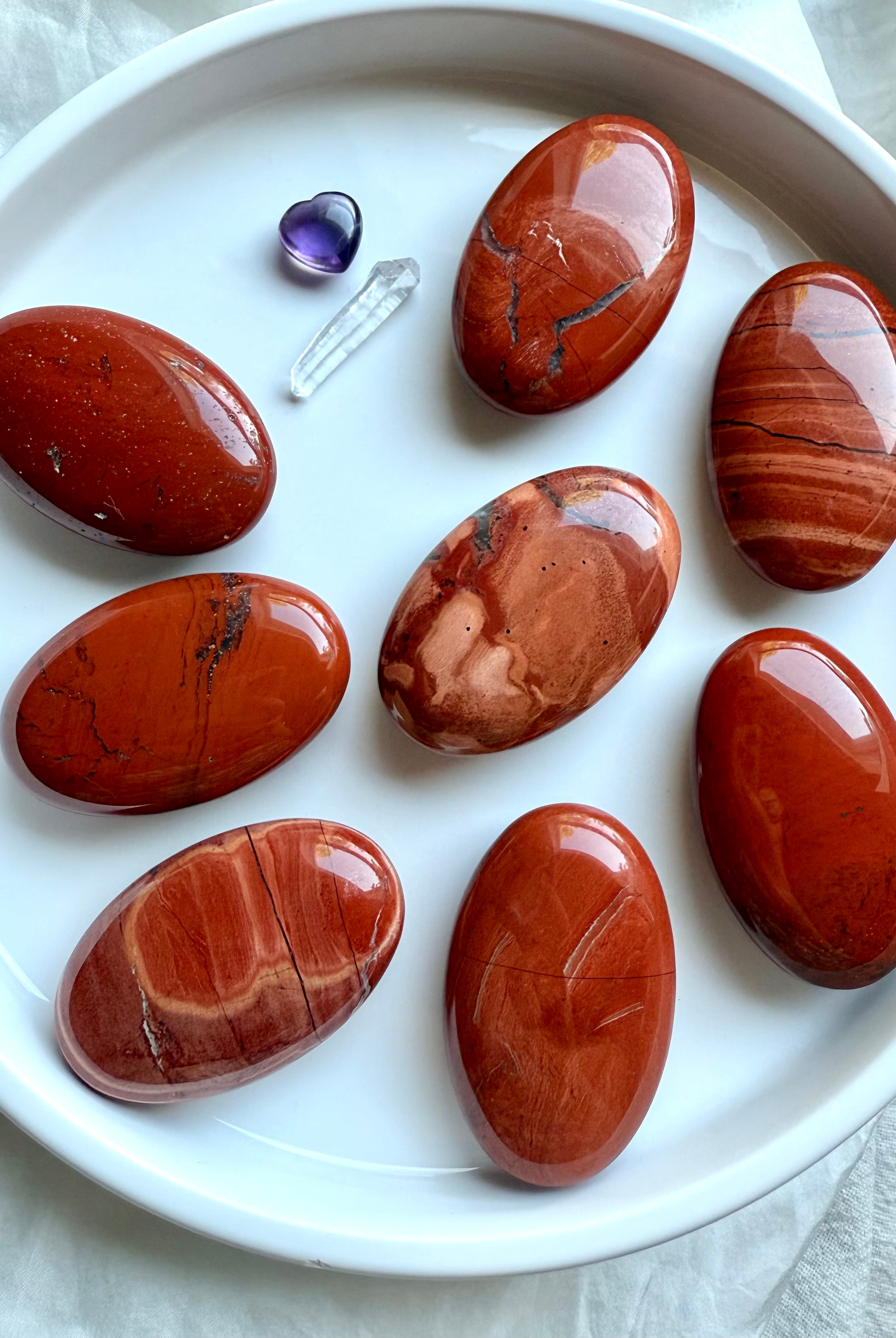 Flatlay of polished Red Jasper palm stones on a white plate — grounding Root Chakra crystals showing natural color and pattern variation.