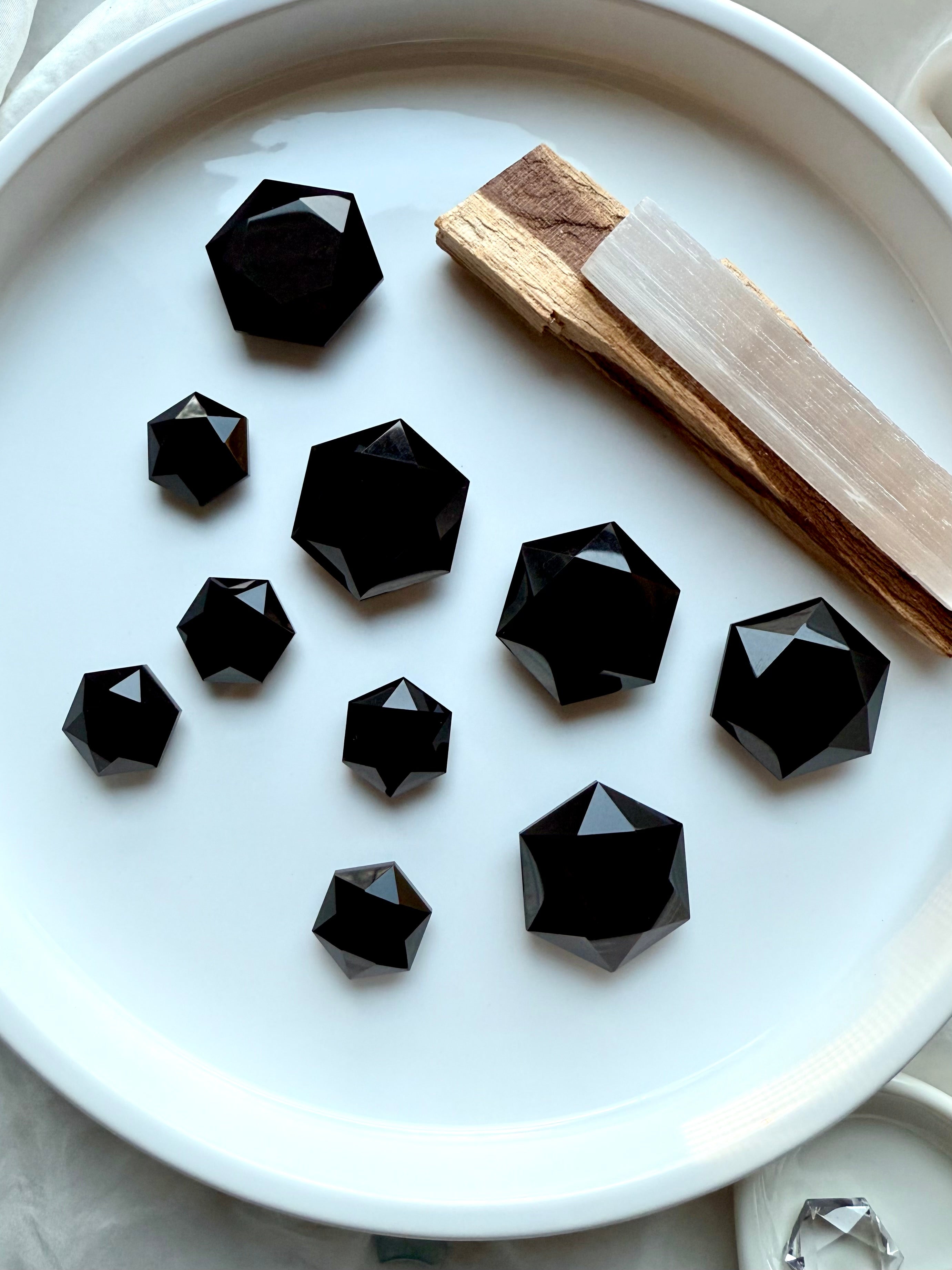 Array of faceted Black Obsidian hexagon crystals on white with selenite and palo santo, showing reflective mirror-like facets and sacred geometry design.