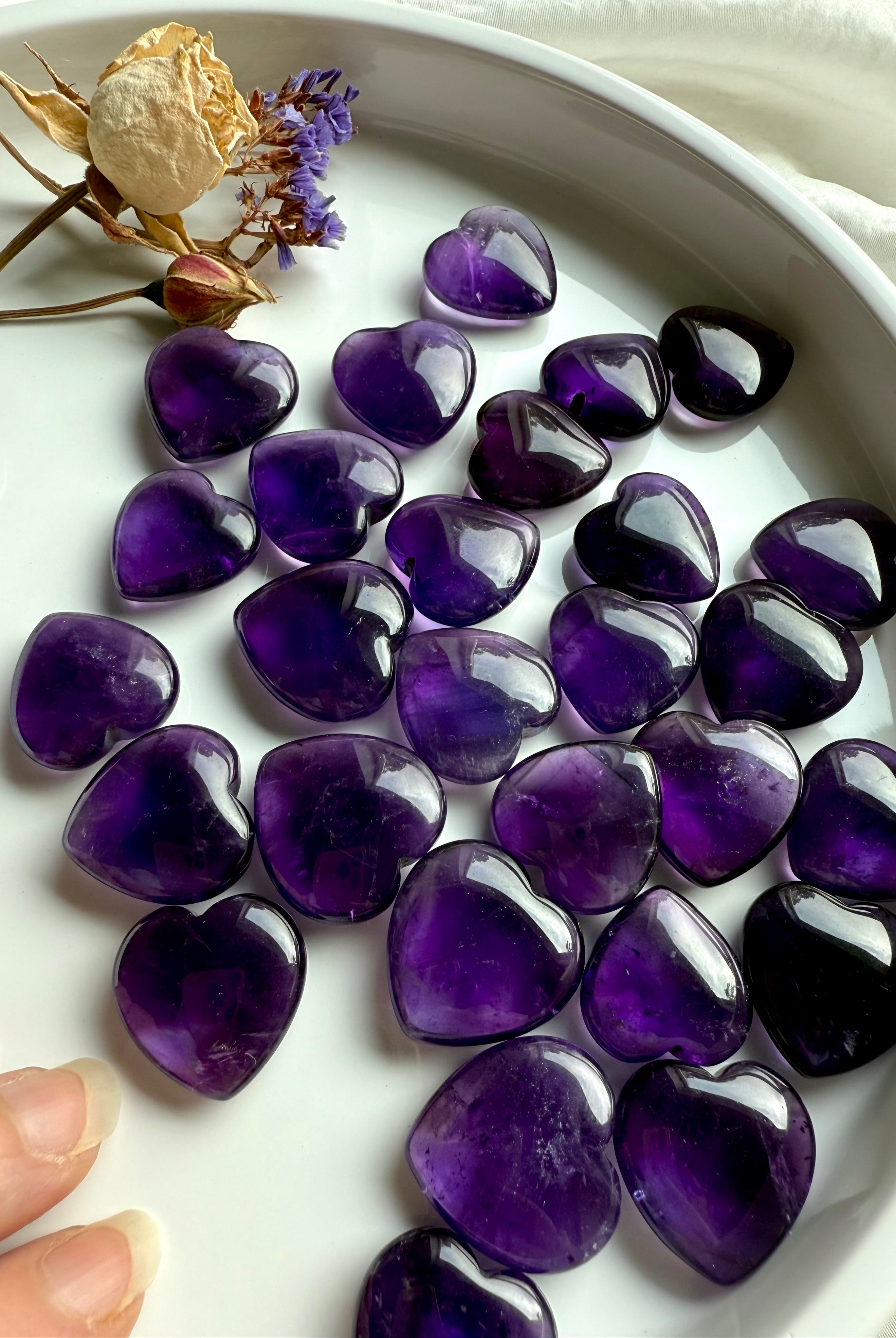 Amethyst crystal hearts in deep purple tones displayed in a dish with dried flowers — polished crown chakra stones for intuition, clarity, and divine guidance