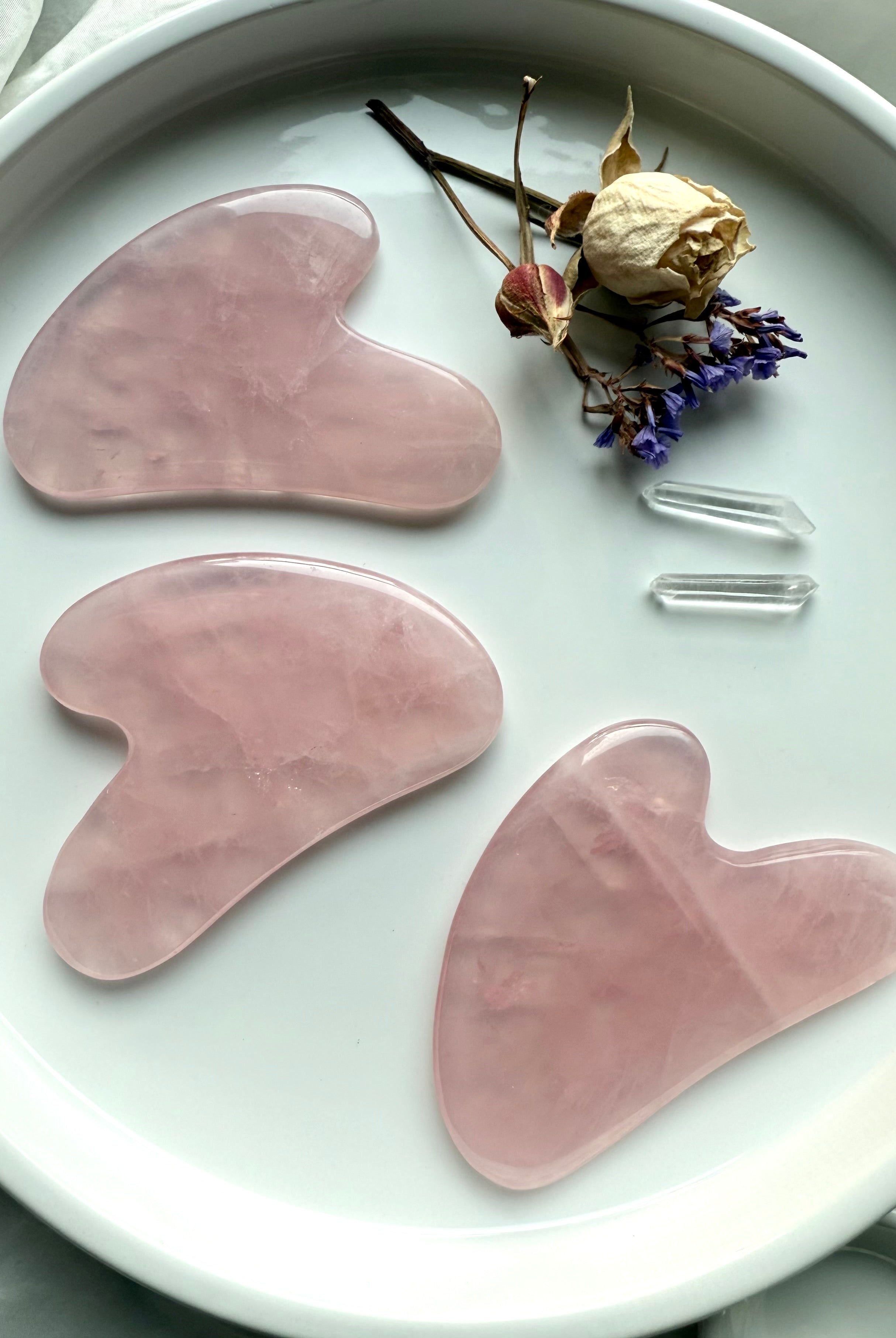Three Rose Quartz crystal gua sha facial massage tools on white plate with flowers and quartz points