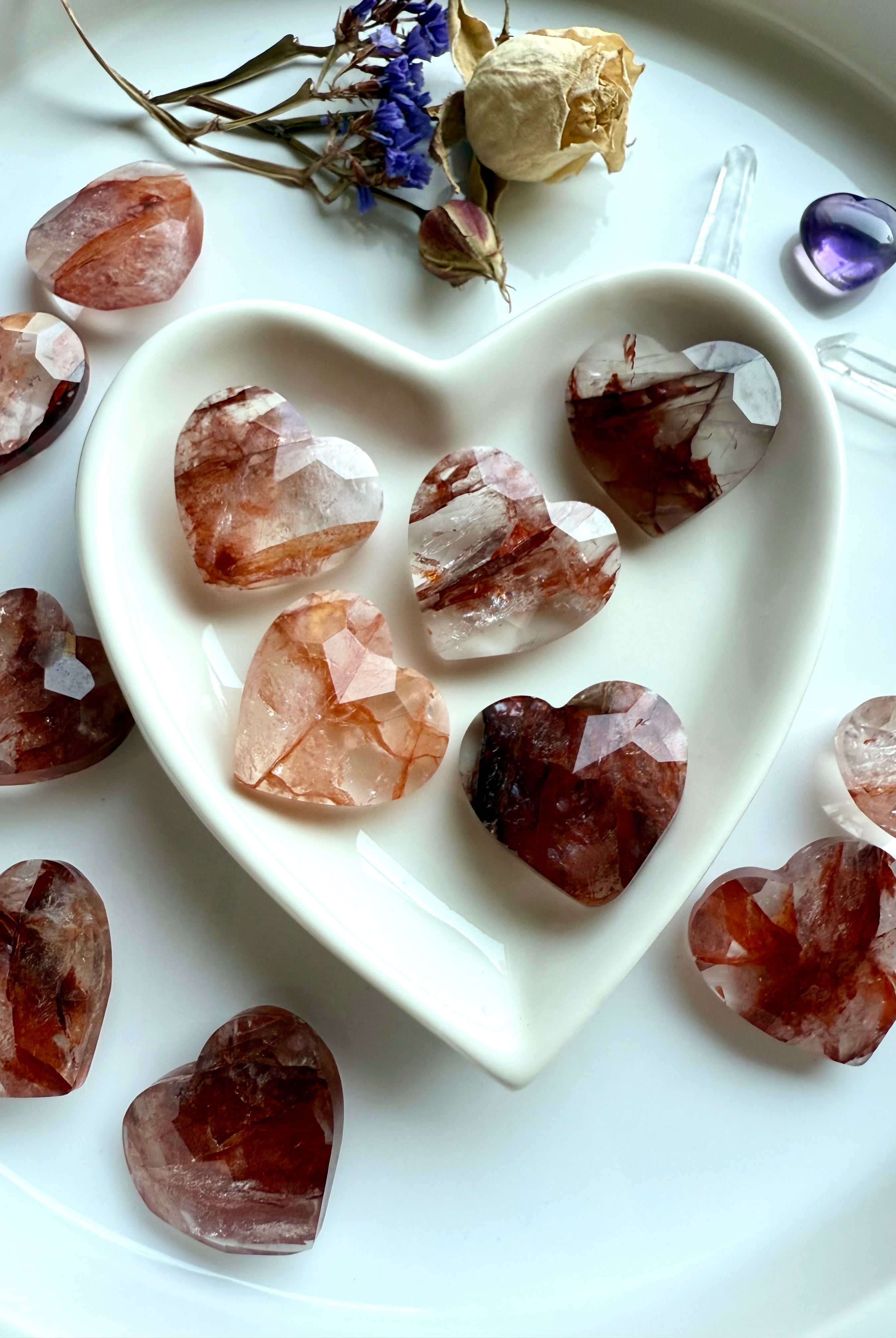 Faceted Fire Quartz crystal hearts in a white heart-shaped bowl with flowers, amethyst heart, and clear quartz points – healing crystals for grounding energy