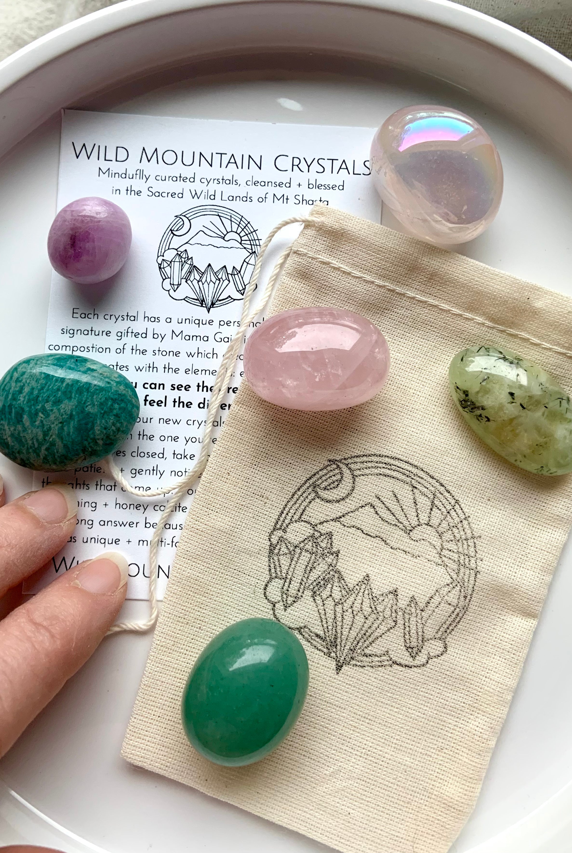 Heart Chakra crystal gift set with 6 pink and green healing stones on top of info card and logo pouch — hand reaching in to select a crystal from the plate.