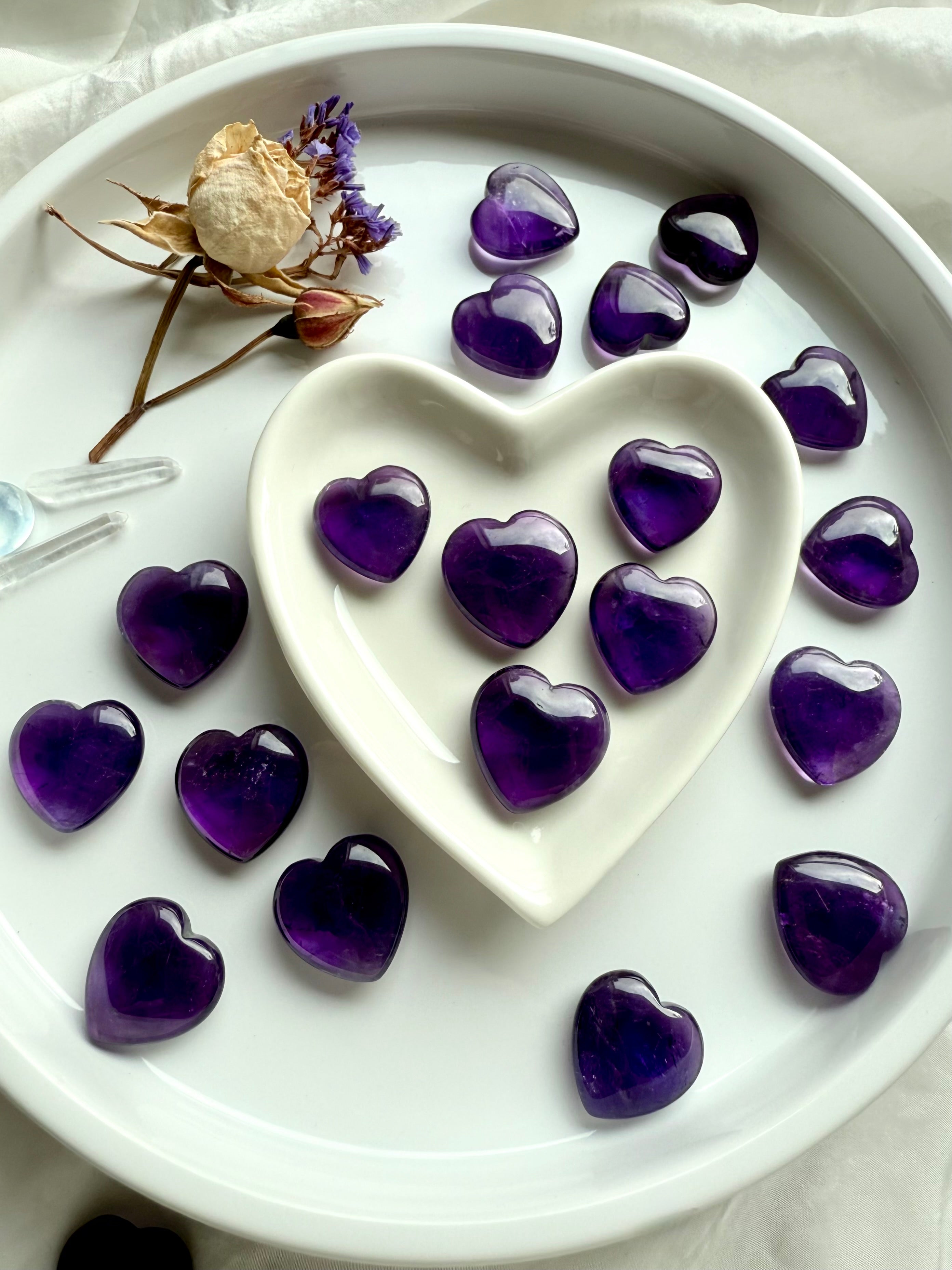 Amethyst crystal hearts displayed around a white heart-shaped bowl with dried flowers — soothing crown chakra crystals for intuition, clarity, and spiritual insight.