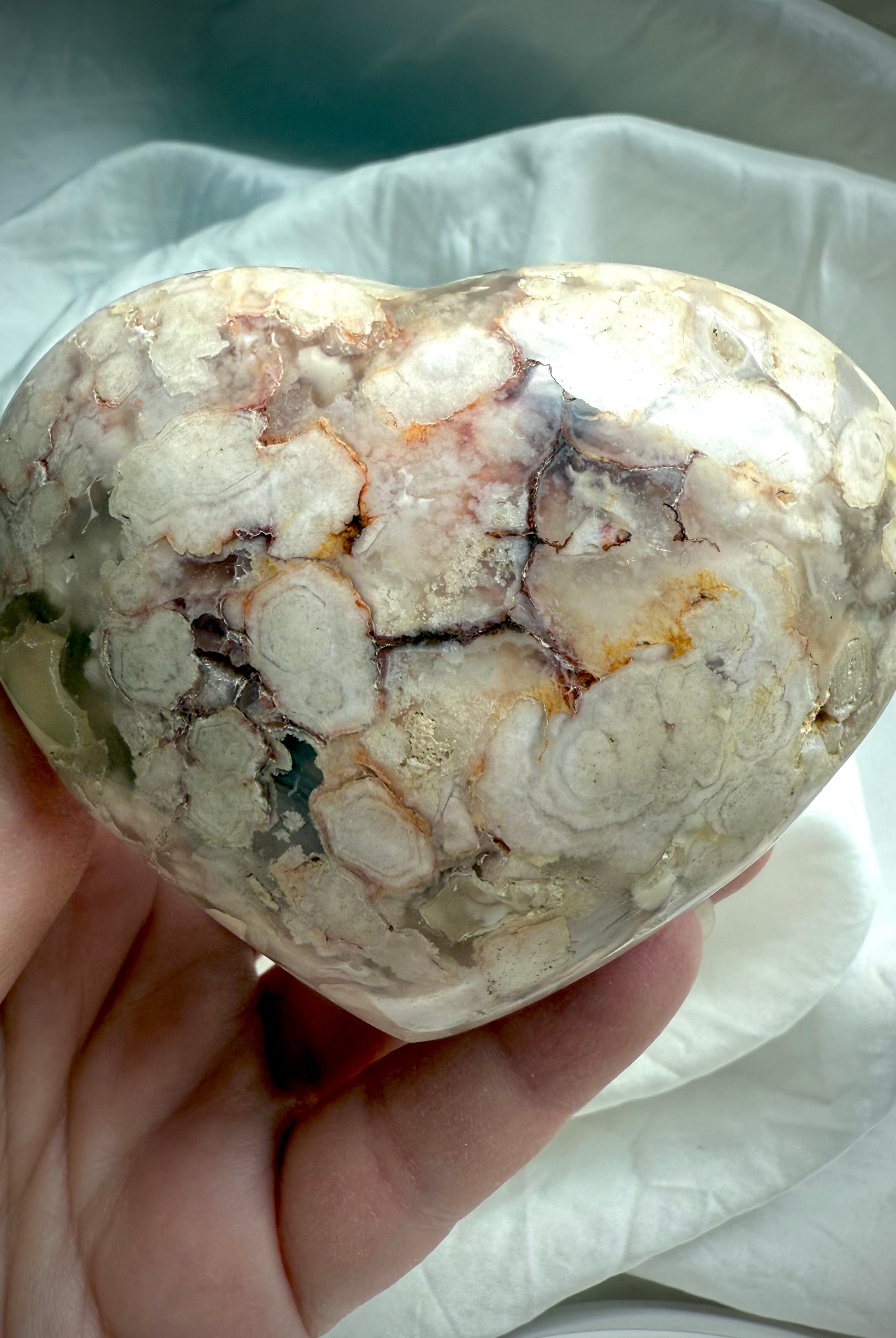 Polished Flower Agate heart resting in an open hand with creamy amber and white blossom formations — supportive stone for comfort, inner growth, and self-love.