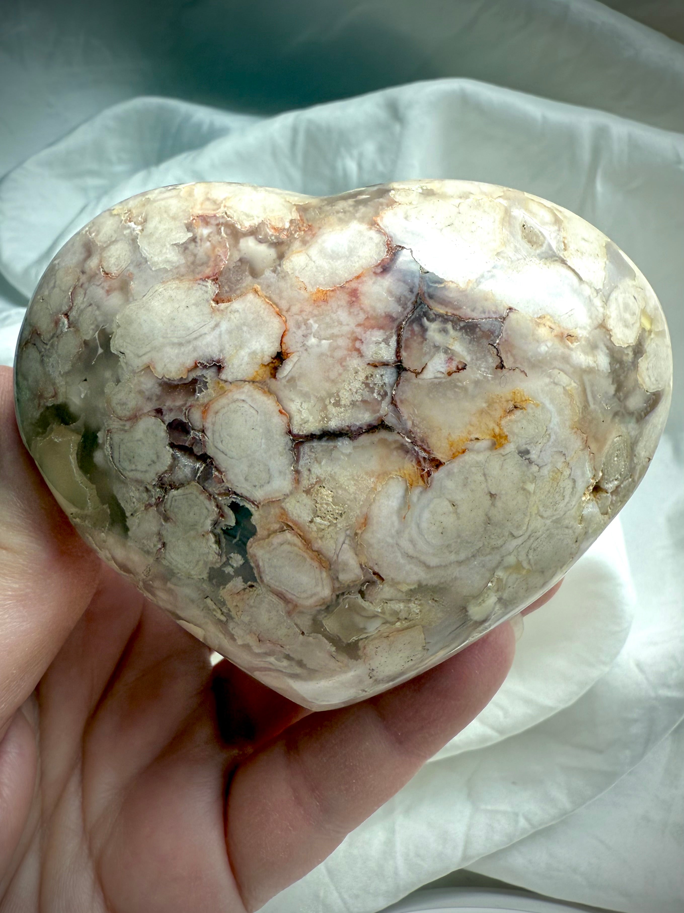 Polished Flower Agate heart resting in an open hand with creamy amber and white blossom formations โ supportive stone for comfort, inner growth, and self-love.