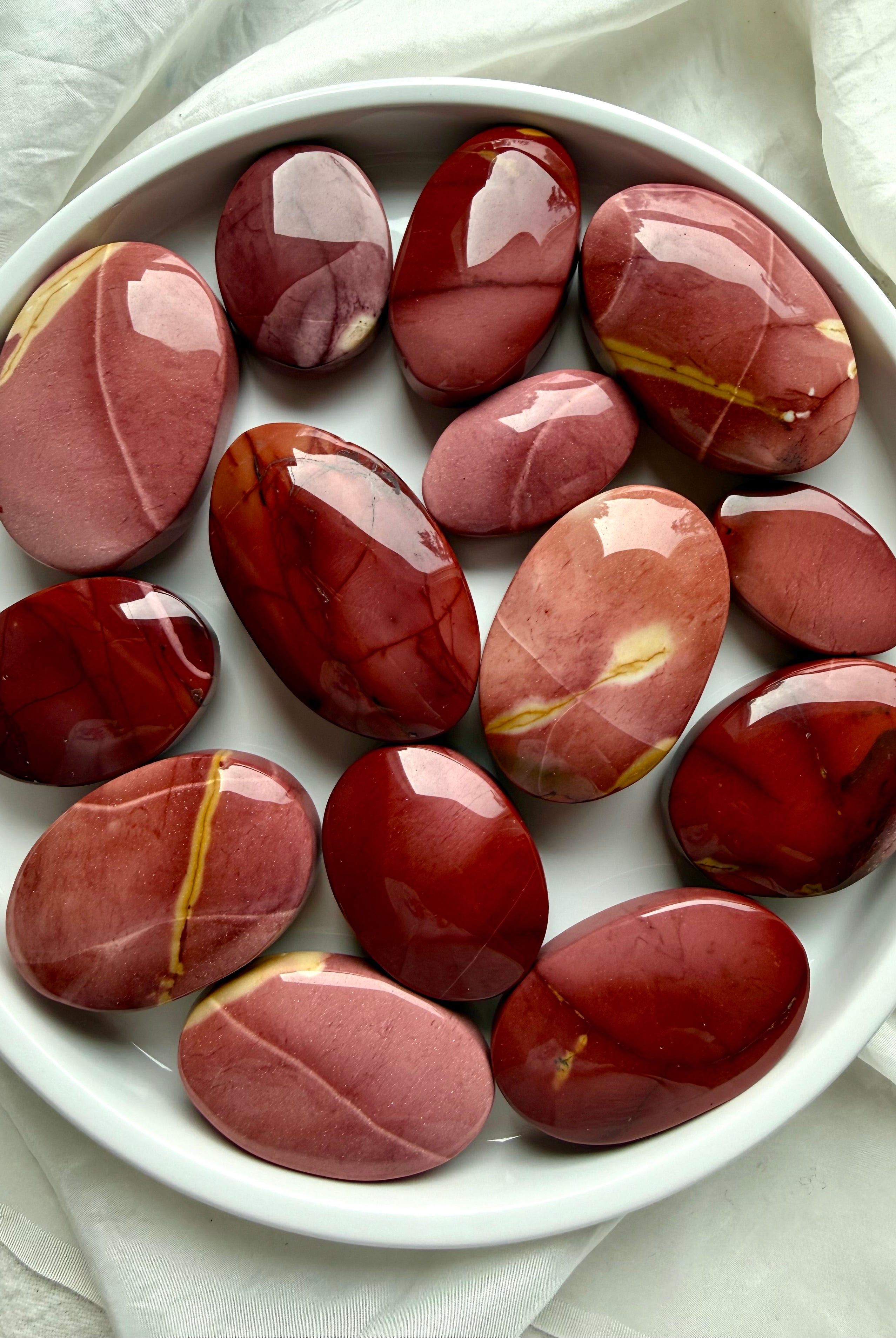 overhead view of polished Mookaite Jasper palm stones on white dish showing earthy red, mauve, cream, and burgundy color variations