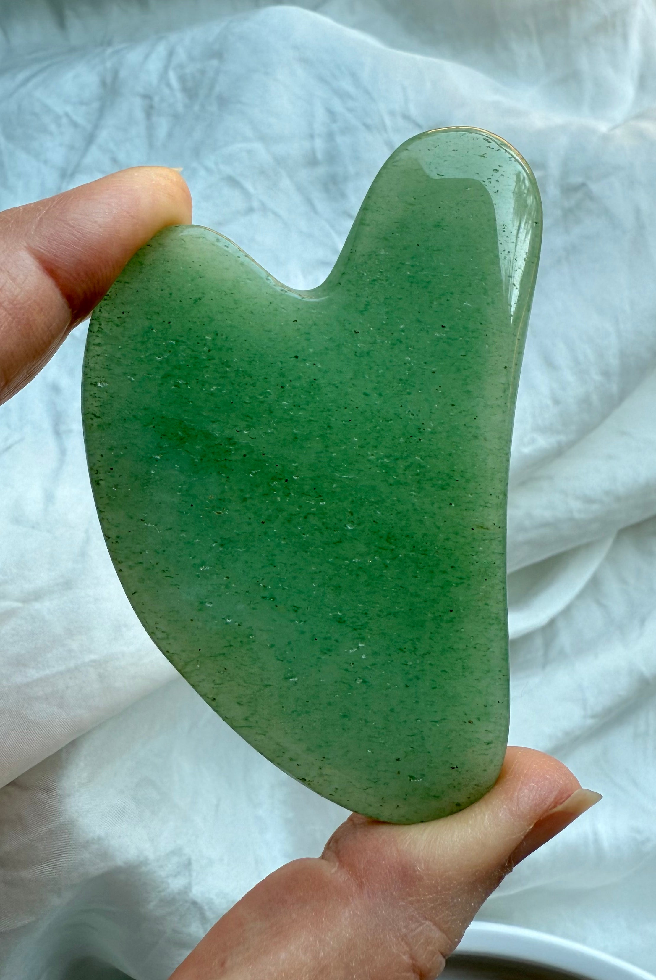 Green Aventurine crystal gua sha facial massage tool held upright against white fabric background