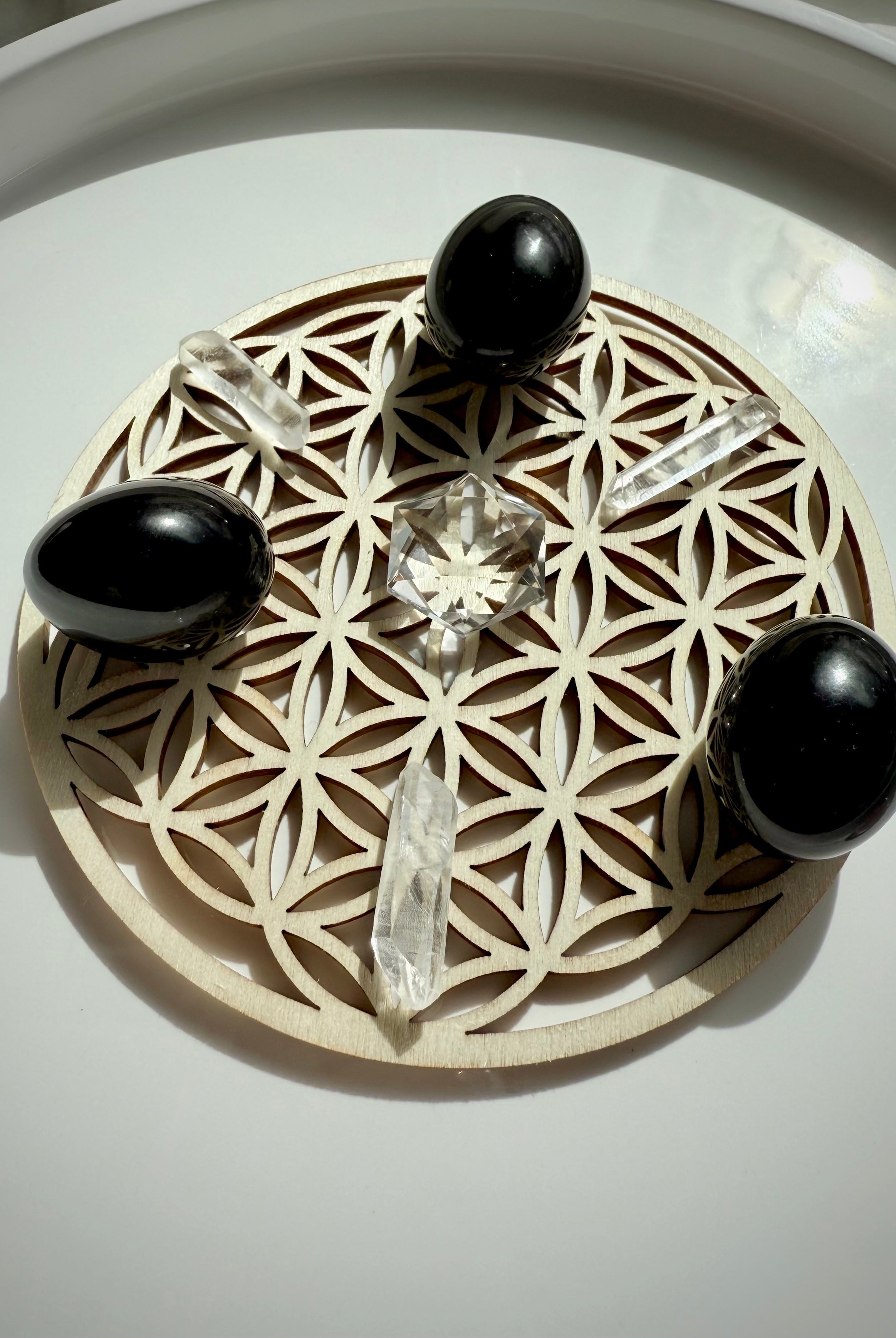 Angled view of crystal grid with small Clear Quartz hexagon centerpiece, arranged with three quartz points and three black obsidian tumbles on a wood Flower of Life grid plate over white surface