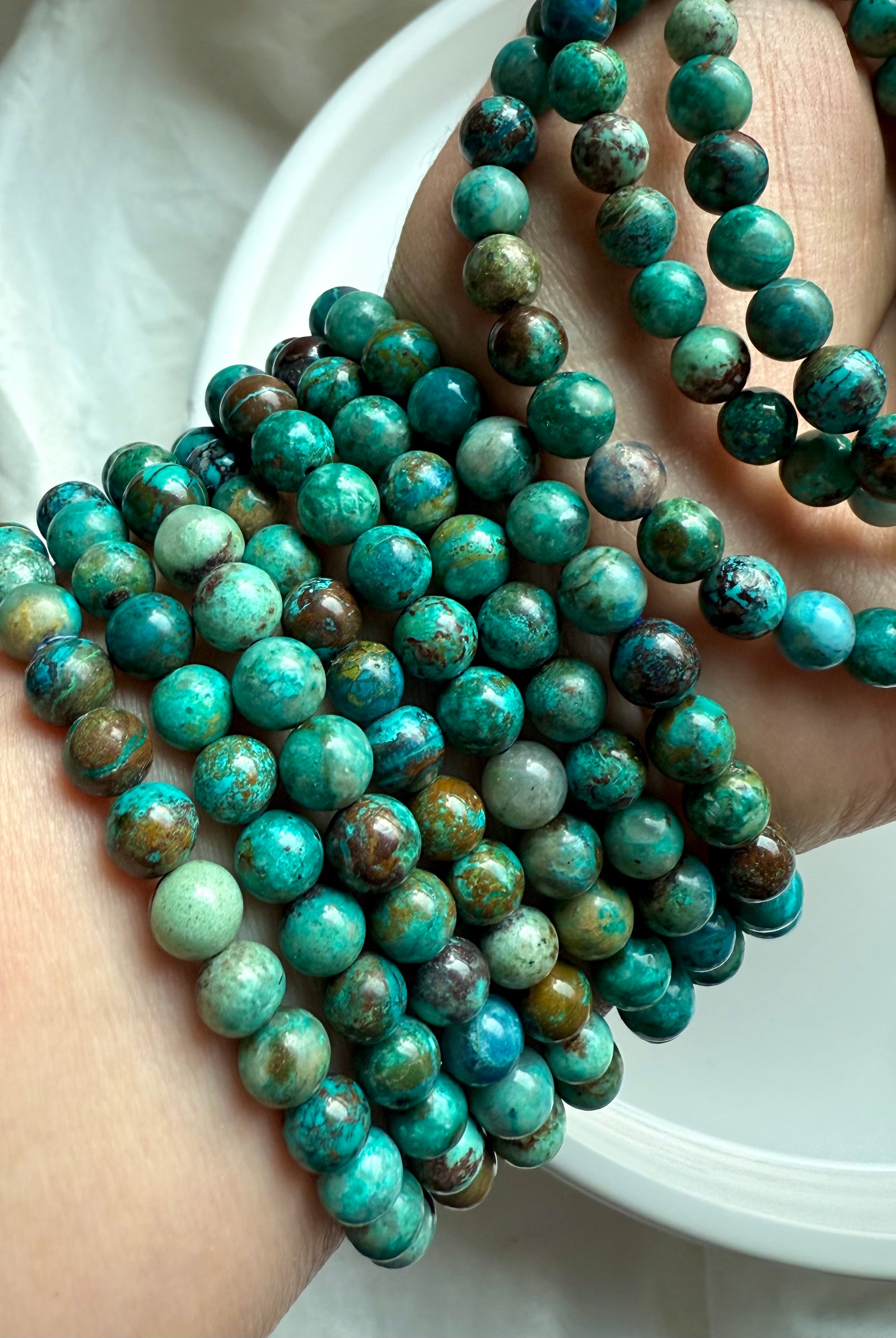 Collection of Chrysocolla Shattuckite stretch bracelets in hand, showing variations of teal, blue, and earthy inclusions - throat chakra healing jewelry