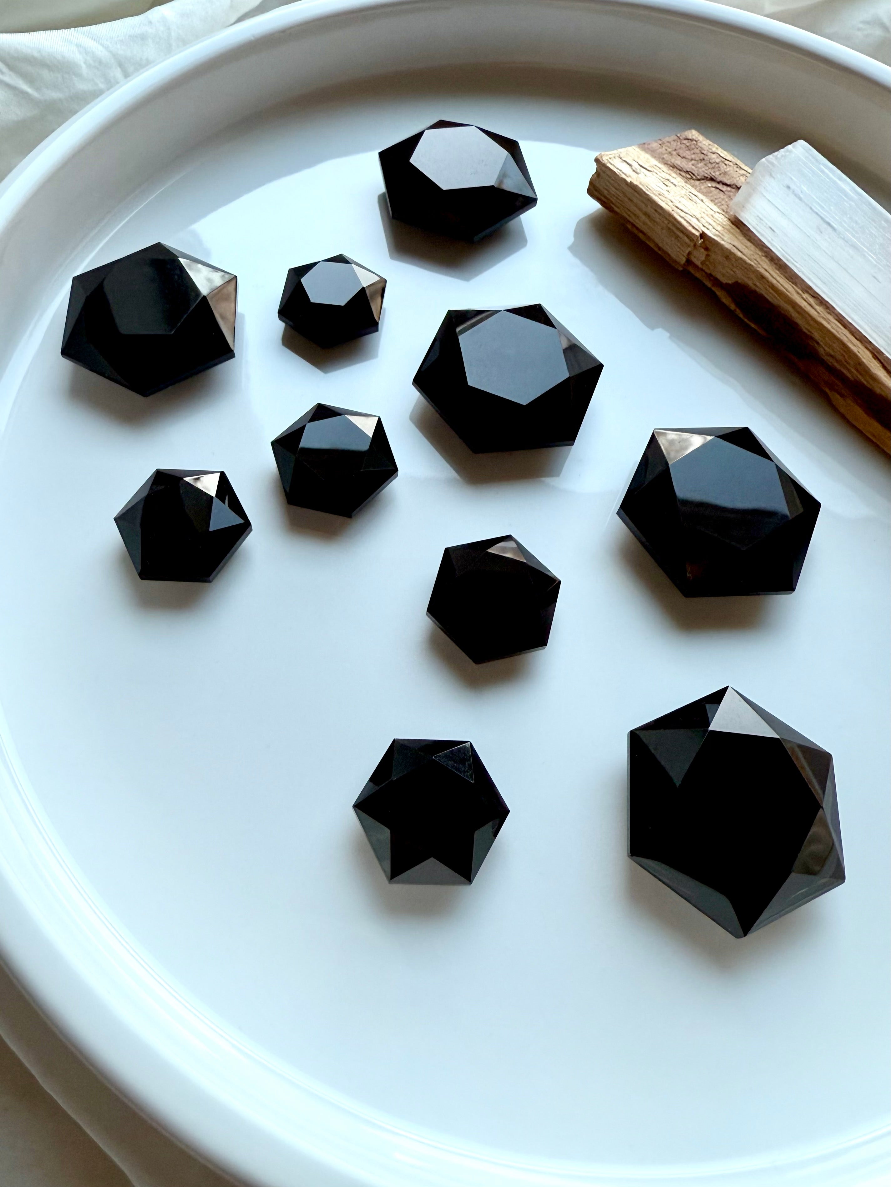 Faceted Black Obsidian hexagon crystals in assorted sizes displayed on a white dish with Palo Santo and Selenite — grounding root chakra stones for protection and energy clearing.