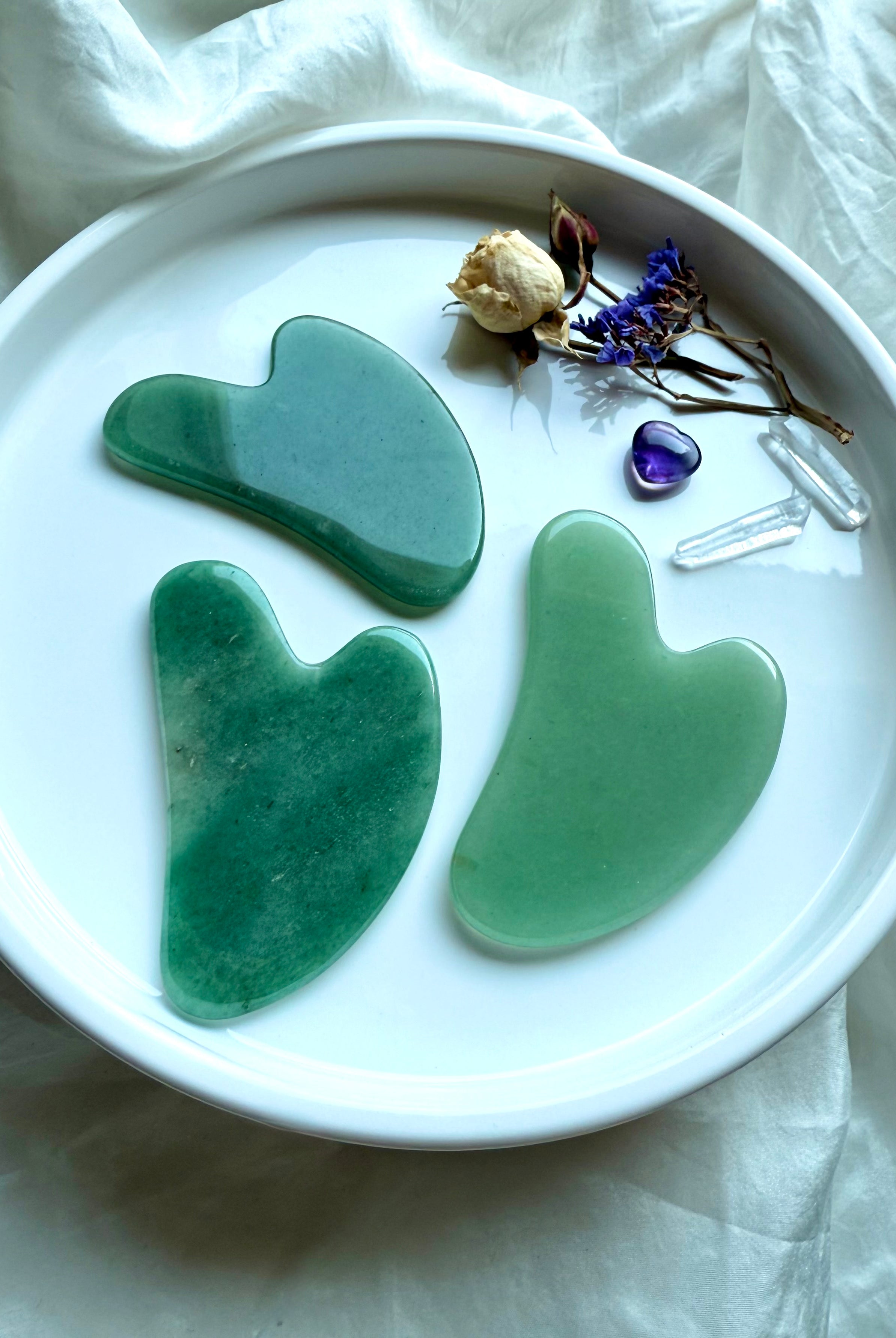 Three Green Aventurine crystal gua sha facial massage tools on white plate with flowers and amethyst stone