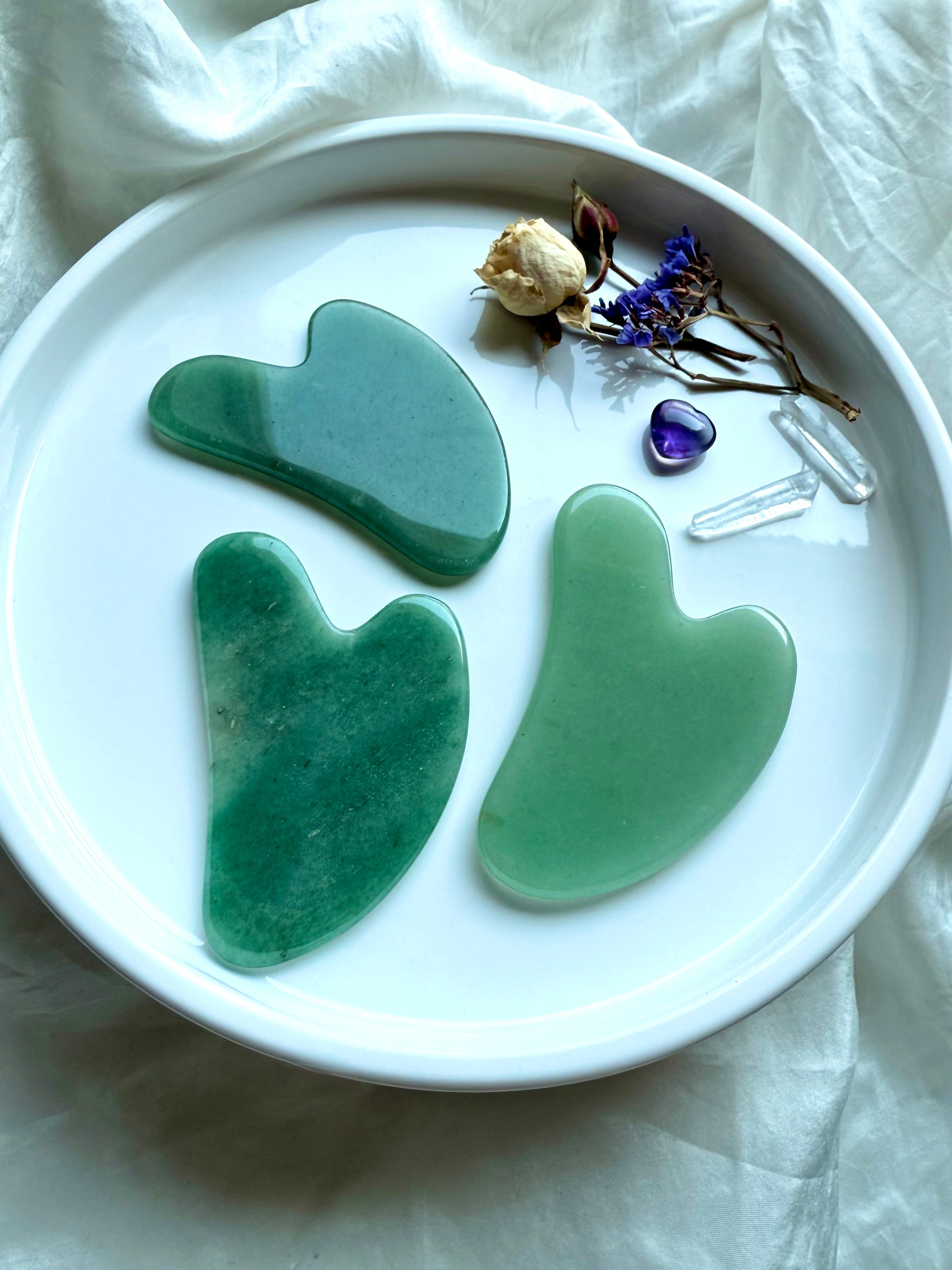 Three Green Aventurine crystal gua sha facial massage tools on white plate with flowers and amethyst stone