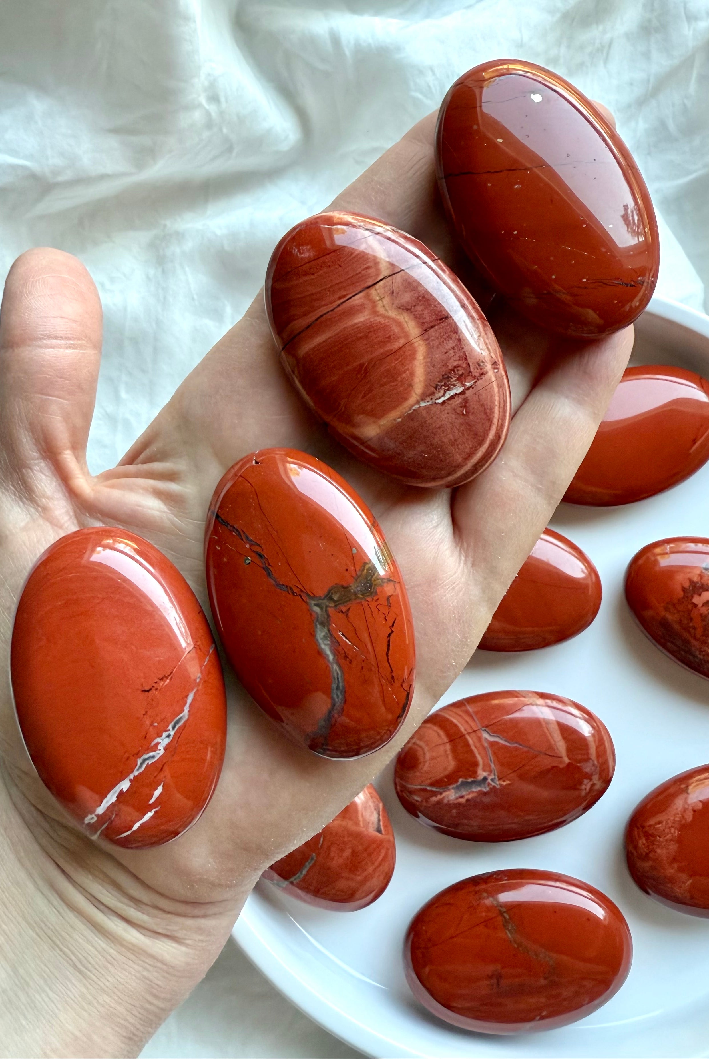 Four Red Jasper palm stones shown on hand above a plate of crystals — earthy healing stones for grounding and strength.
