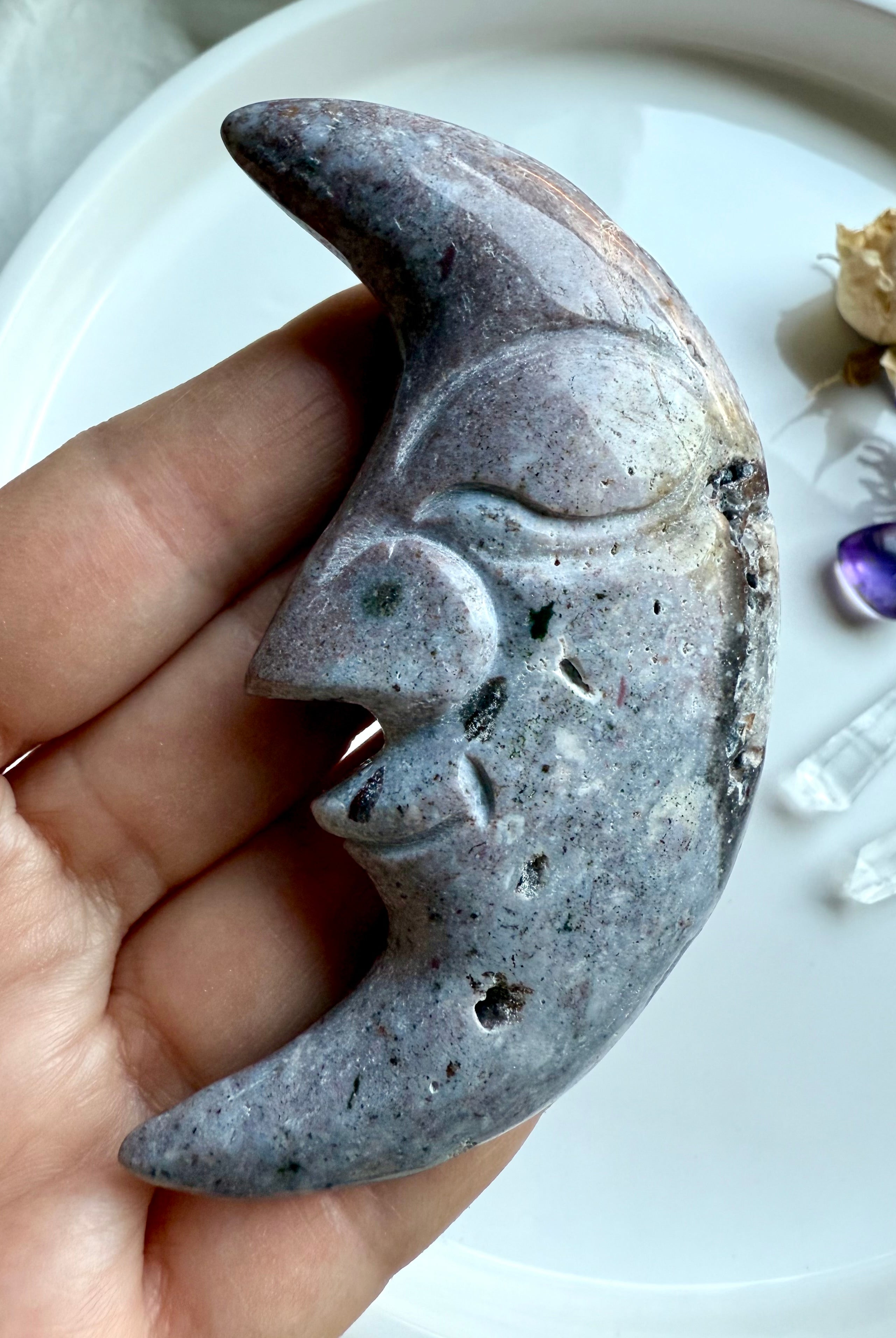 Ocean Jasper moon face carving in purple tones styled with flowers and healing crystals for decorative display.
