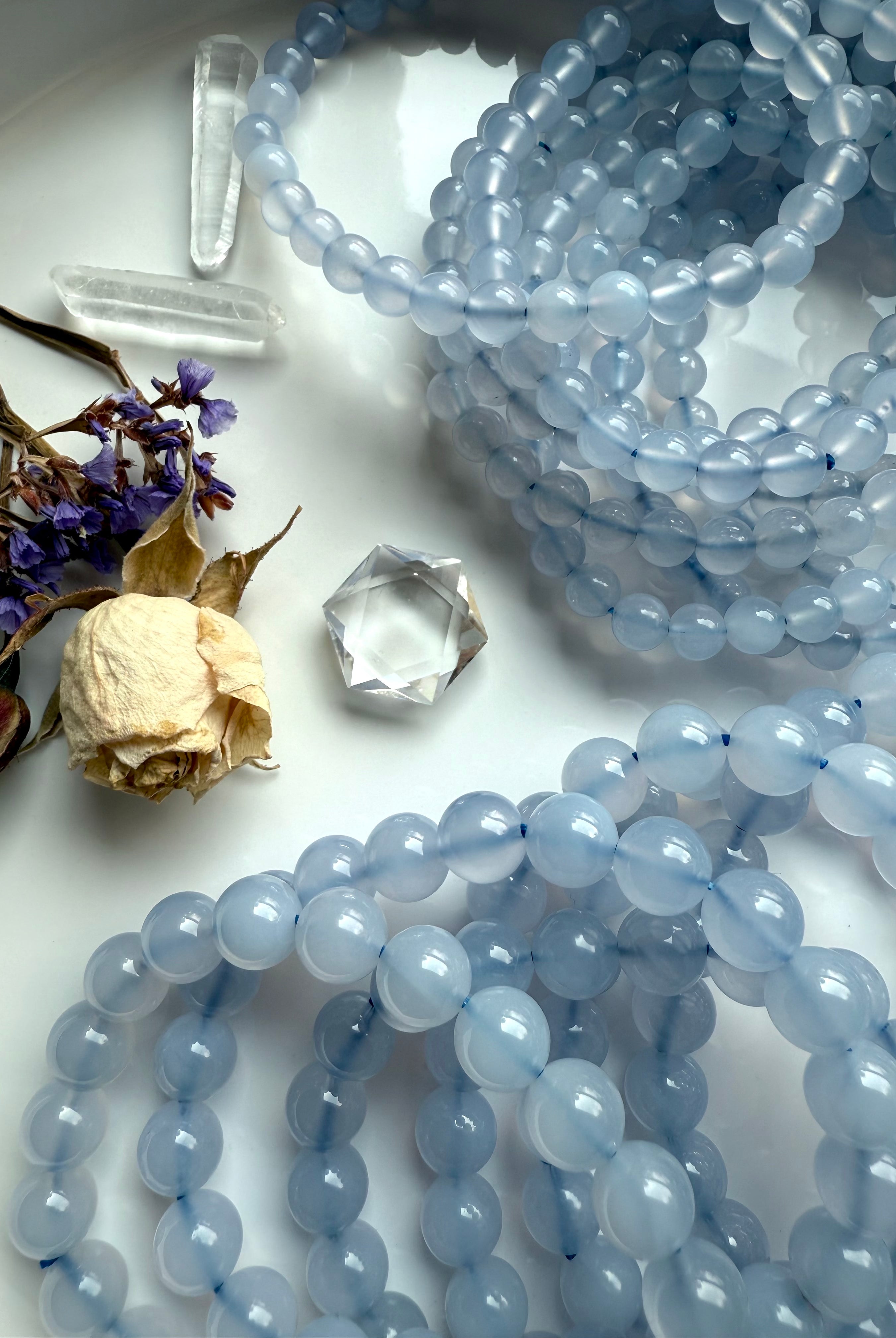 Blue Chalcedony bead bracelets displayed in gentle coils on a white ceramic plate with dried flowers — soothing crystal jewelry for calm and clarity.