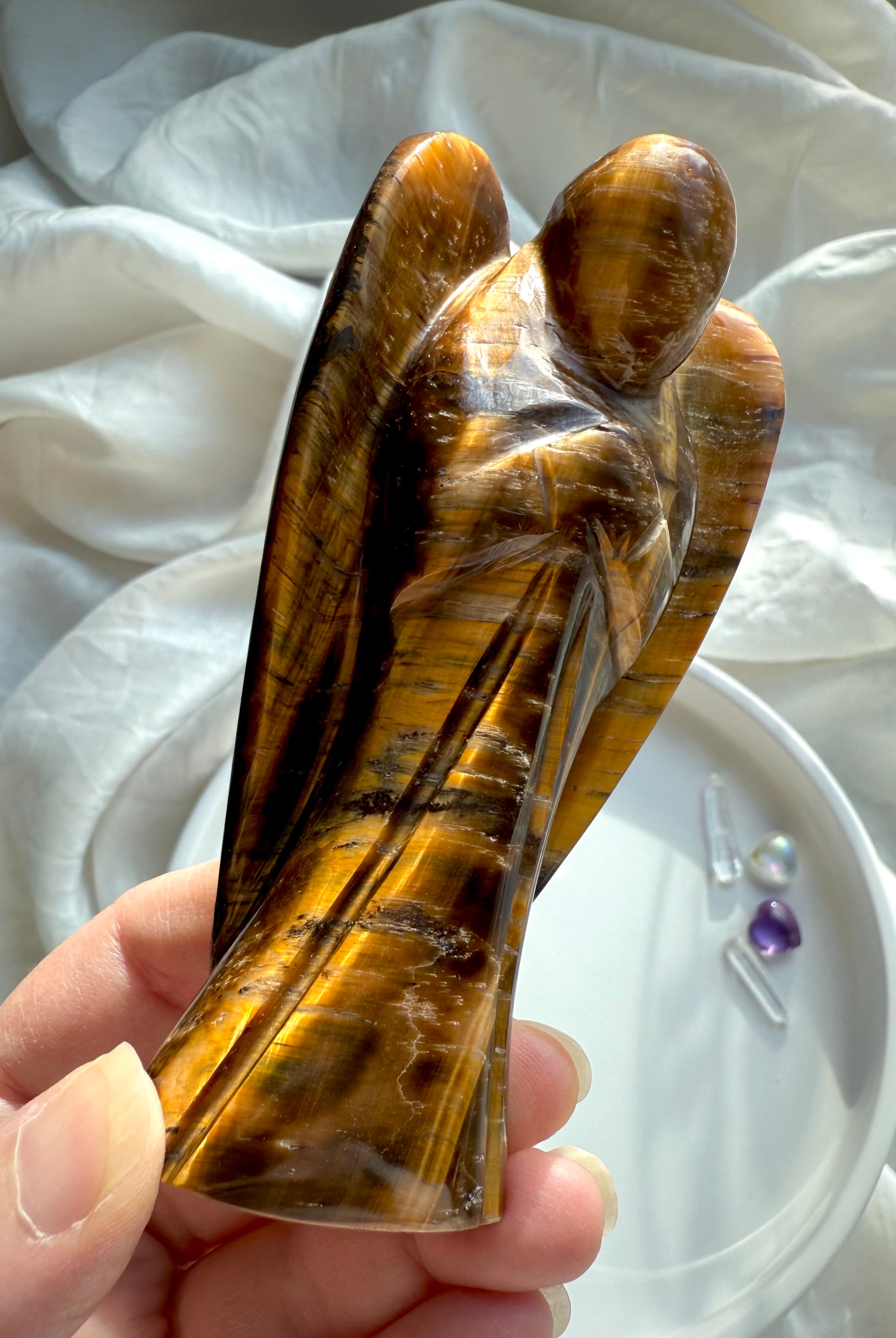 Side view of Tiger’s Eye crystal angel carving glowing in natural light, showcasing chatoyant bands and protective wings