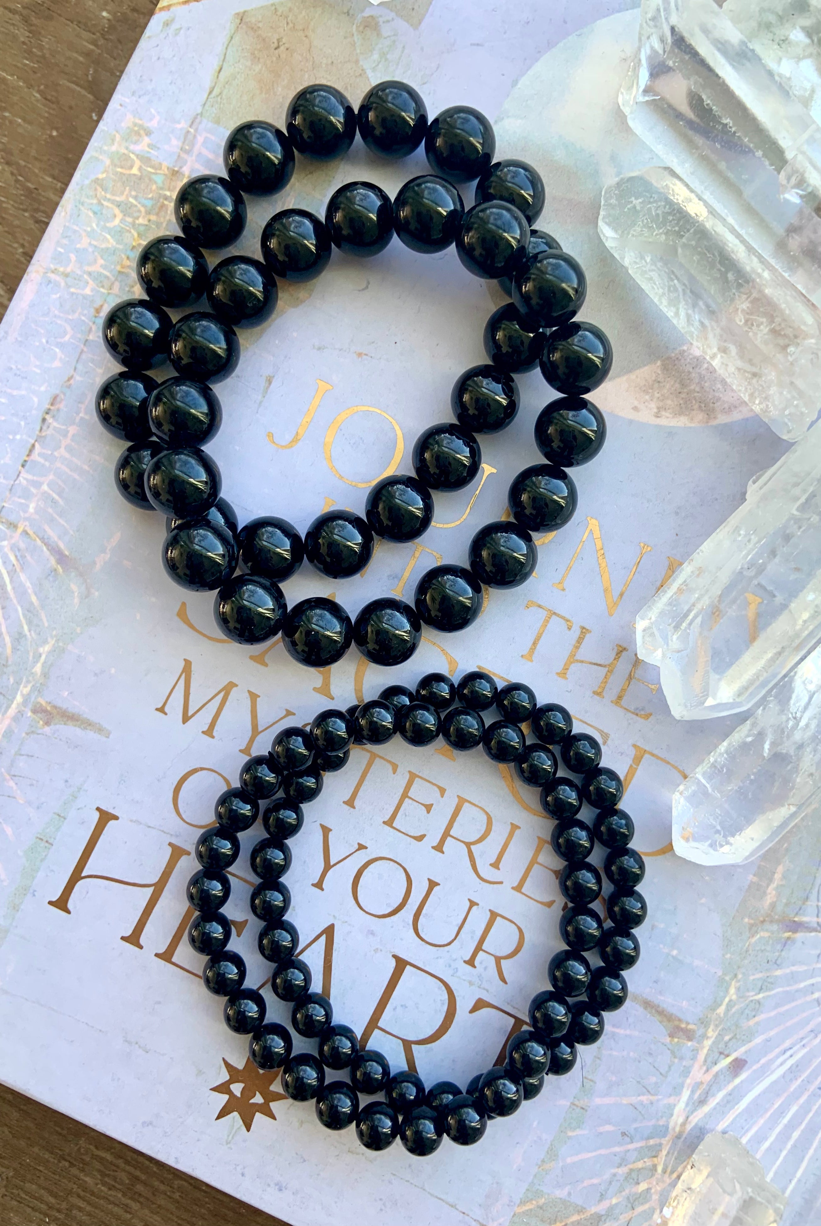 Two sizes of Black Tourmaline bracelets next to clear quartz points in a flatlay — glossy 10–11mm and 6–7mm bead sizes shown for comparison