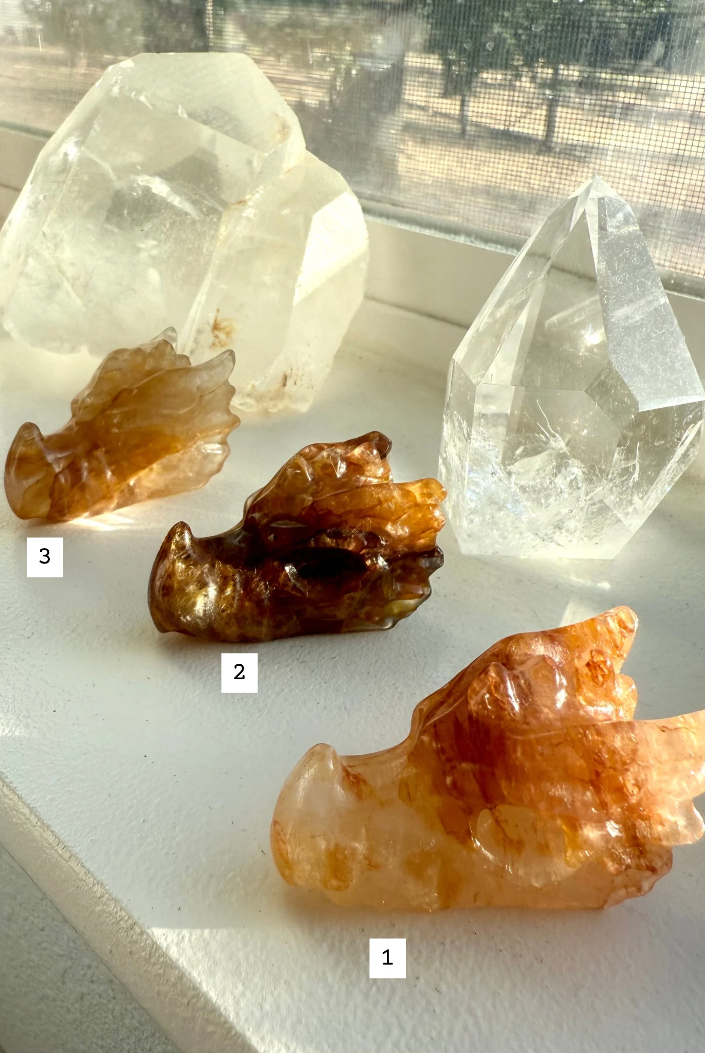 Numbered Golden Healer Quartz dragon head carvings displayed on windowsill — crystal dragon figurines with golden hematite inclusions for chakra healing and spiritual gifting.