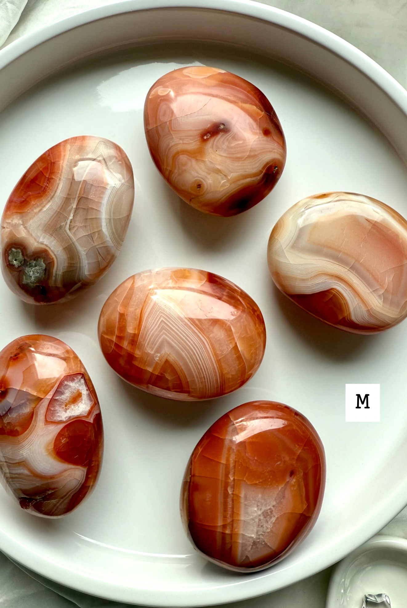Medium Carnelian palm stones with rich red-orange banding on a white plate — grounding crystals for creativity, confidence + life force energy.