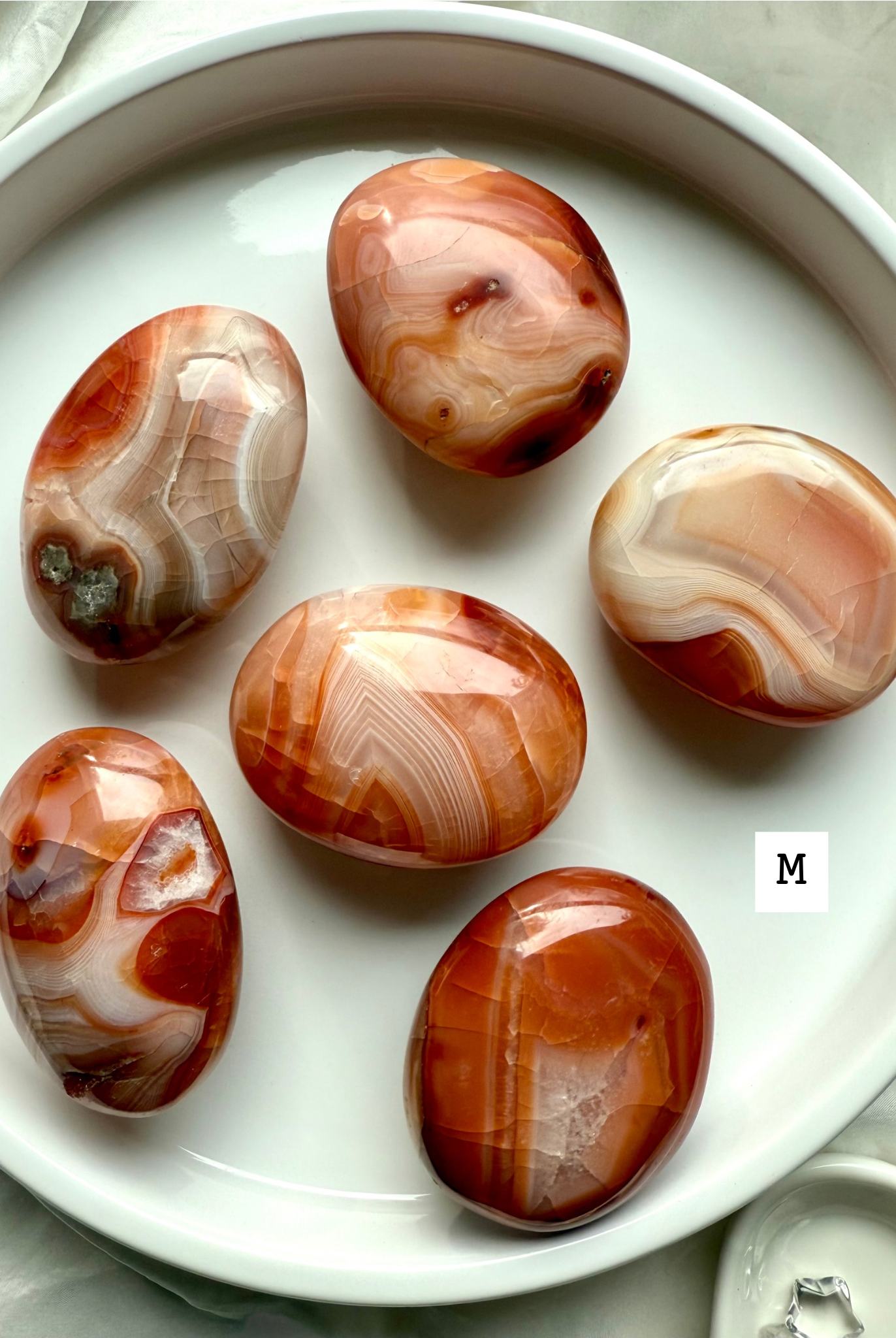 Medium Carnelian palm stones with rich red-orange banding on a white plate — grounding crystals for creativity, confidence + life force energy.