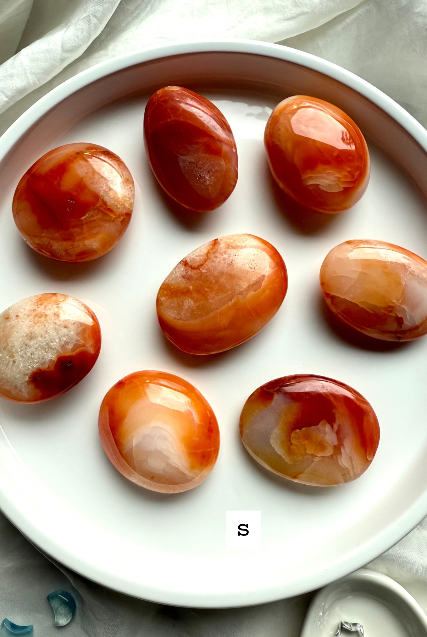 Polished small Carnelian palm stones on a white dish labeled size S — orange and red banded agate crystals for vitality, motivation + Sacral Chakra healing.
