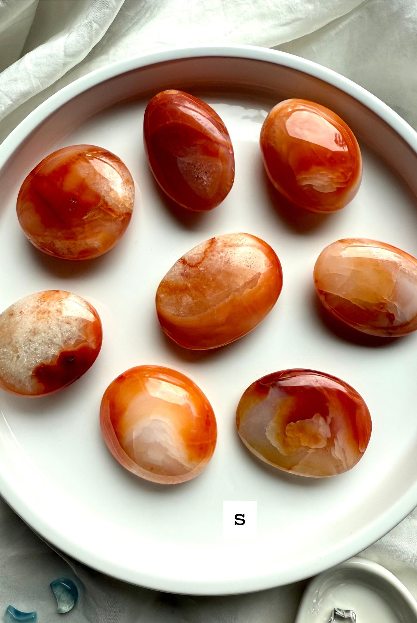 Polished small Carnelian palm stones on a white dish labeled size S — orange and red banded agate crystals for vitality, motivation + Sacral Chakra healing.
