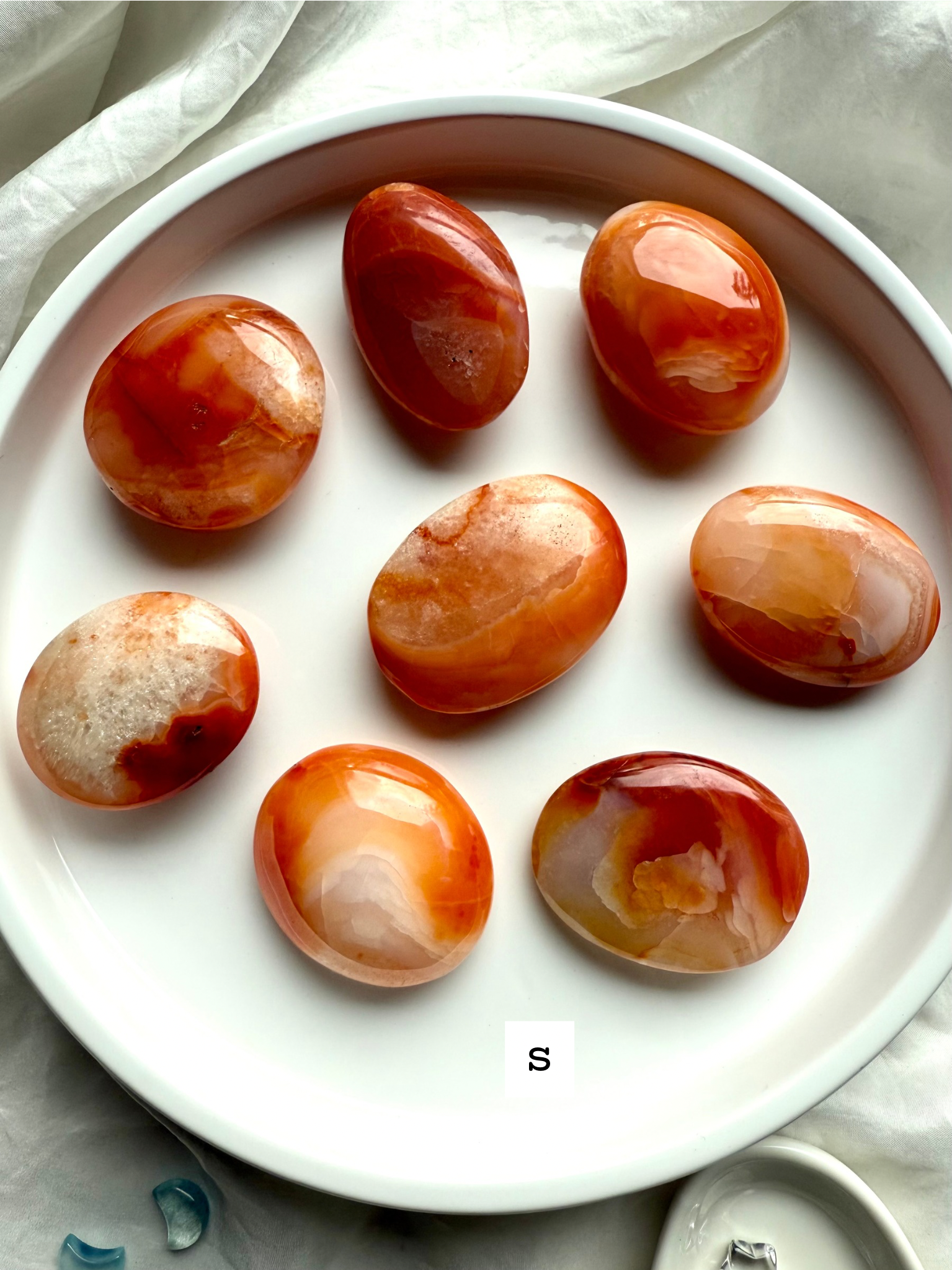 Polished small Carnelian palm stones on a white dish labeled size S — orange and red banded agate crystals for vitality, motivation + Sacral Chakra healing.