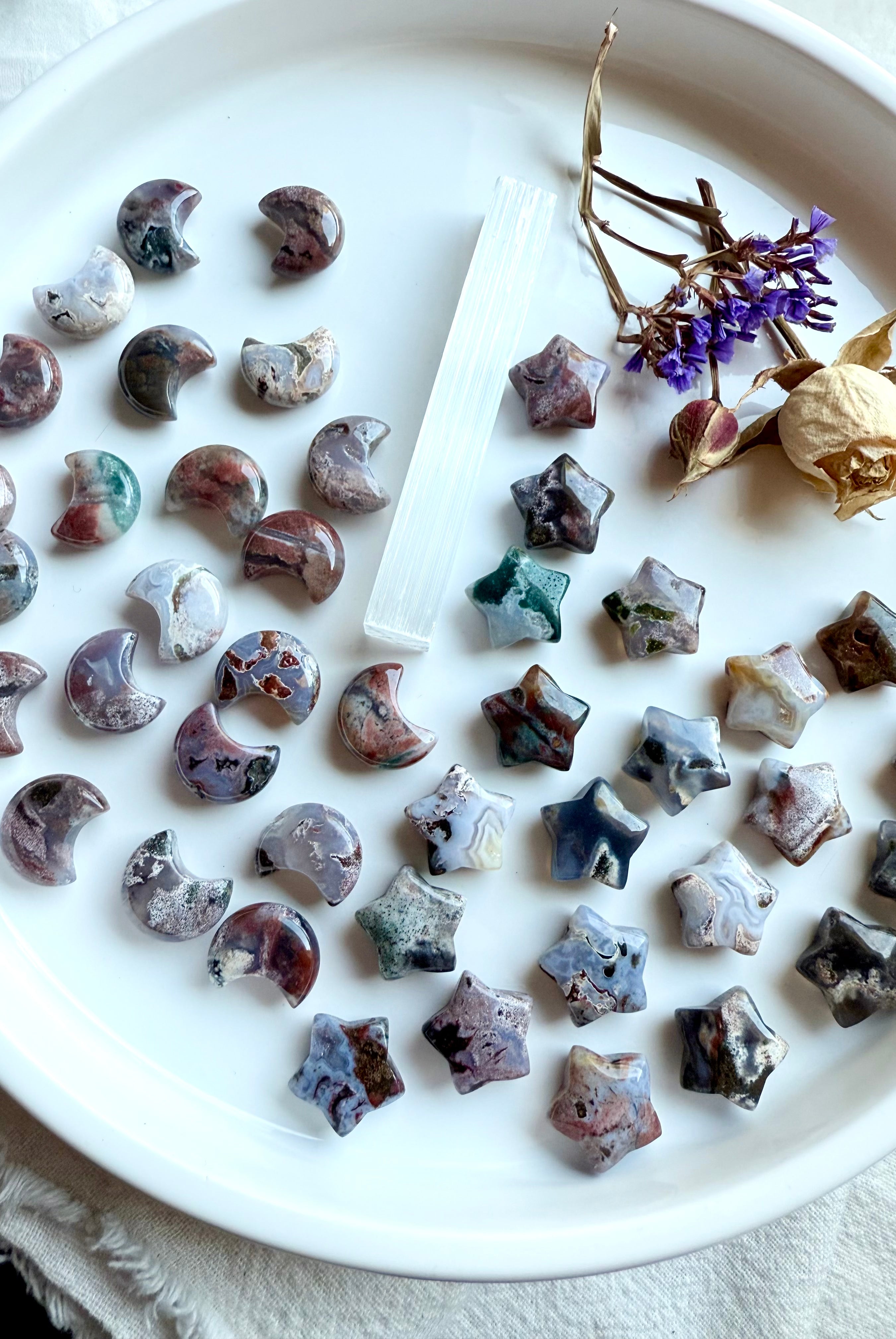 Mini Cosmic Ocean Jasper moons and stars — hand-carved crystal carvings in earthy tones with swirling cosmic patterns on a white dish with flowers and selenite