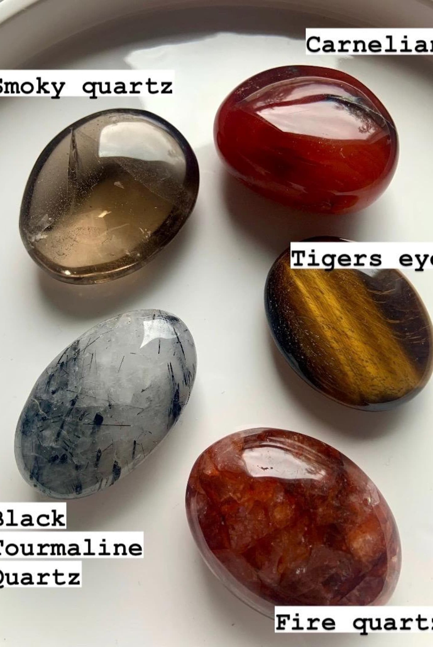 Labeled chakra palm stones on white plate — high-quality polished crystals for lower chakra healing: Smokey Quartz, Tigers Eye, Carnelian, Fire Quartz & Tourmaline in Quartz.