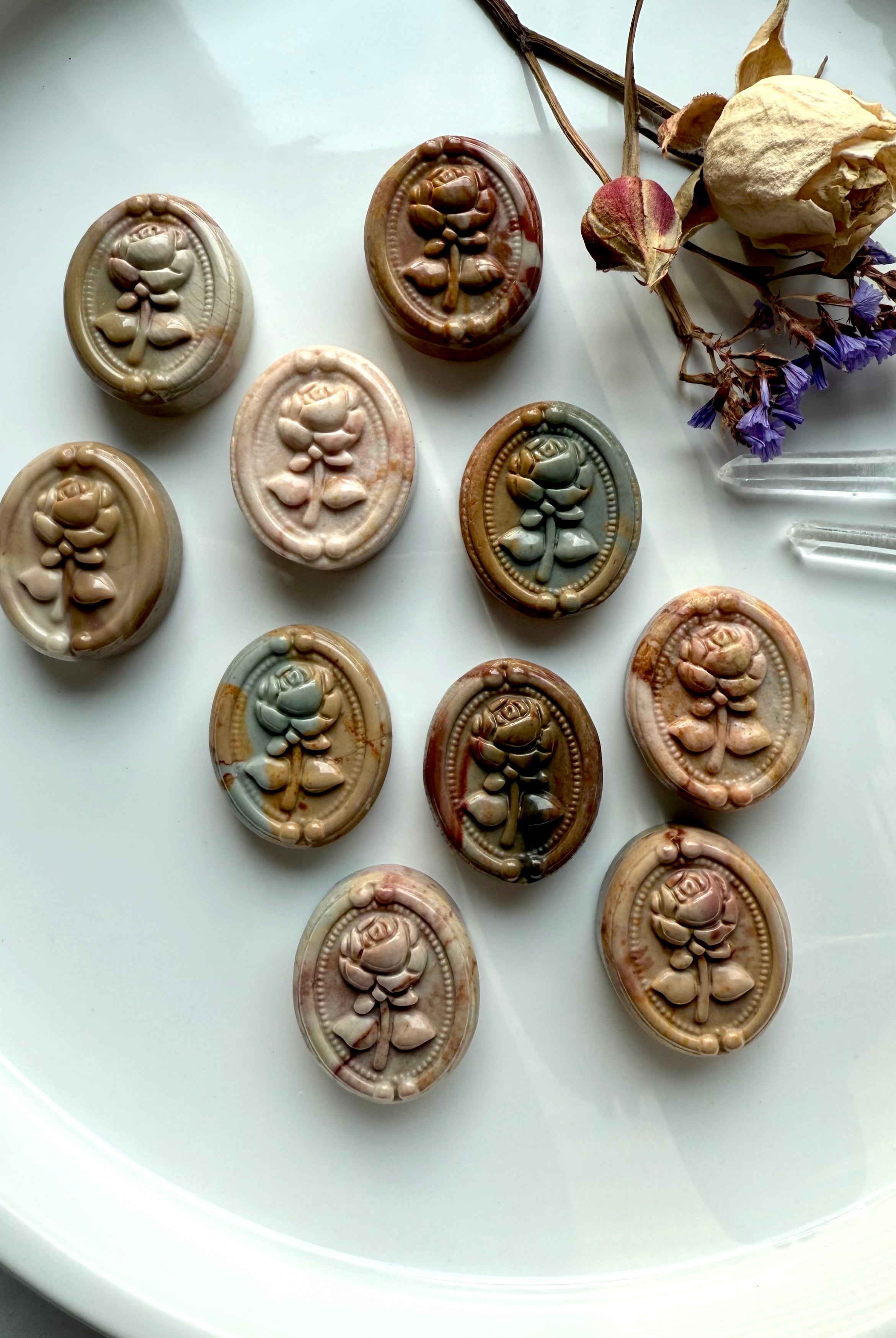 Polychrome Jasper rose cabochons displayed in a group of ten on a white dish with dried flowers and clear quartz points — grounding earth-energy crystals for balance and stability.