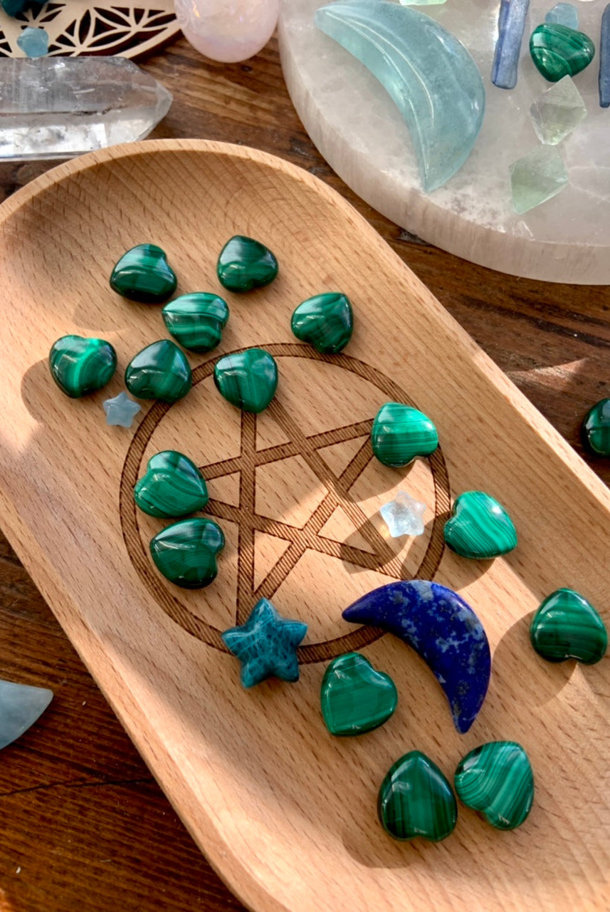 Malachite hearts arranged on a wooden tray with a pentacle engraving, alongside celestial carvings of blue and aqua crystals.
