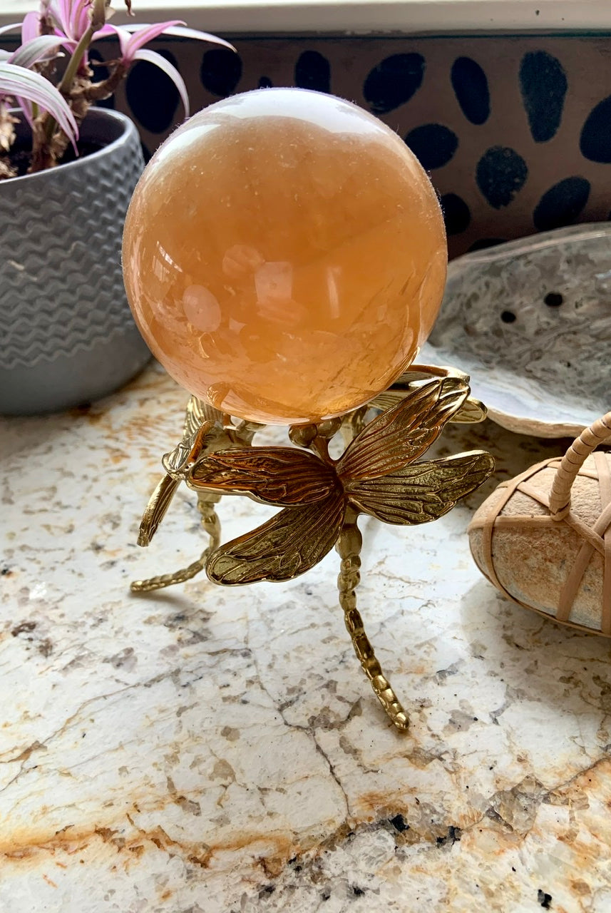 Side view of gold dragonfly crystal stand holding a honey calcite sphere on a marble surface.